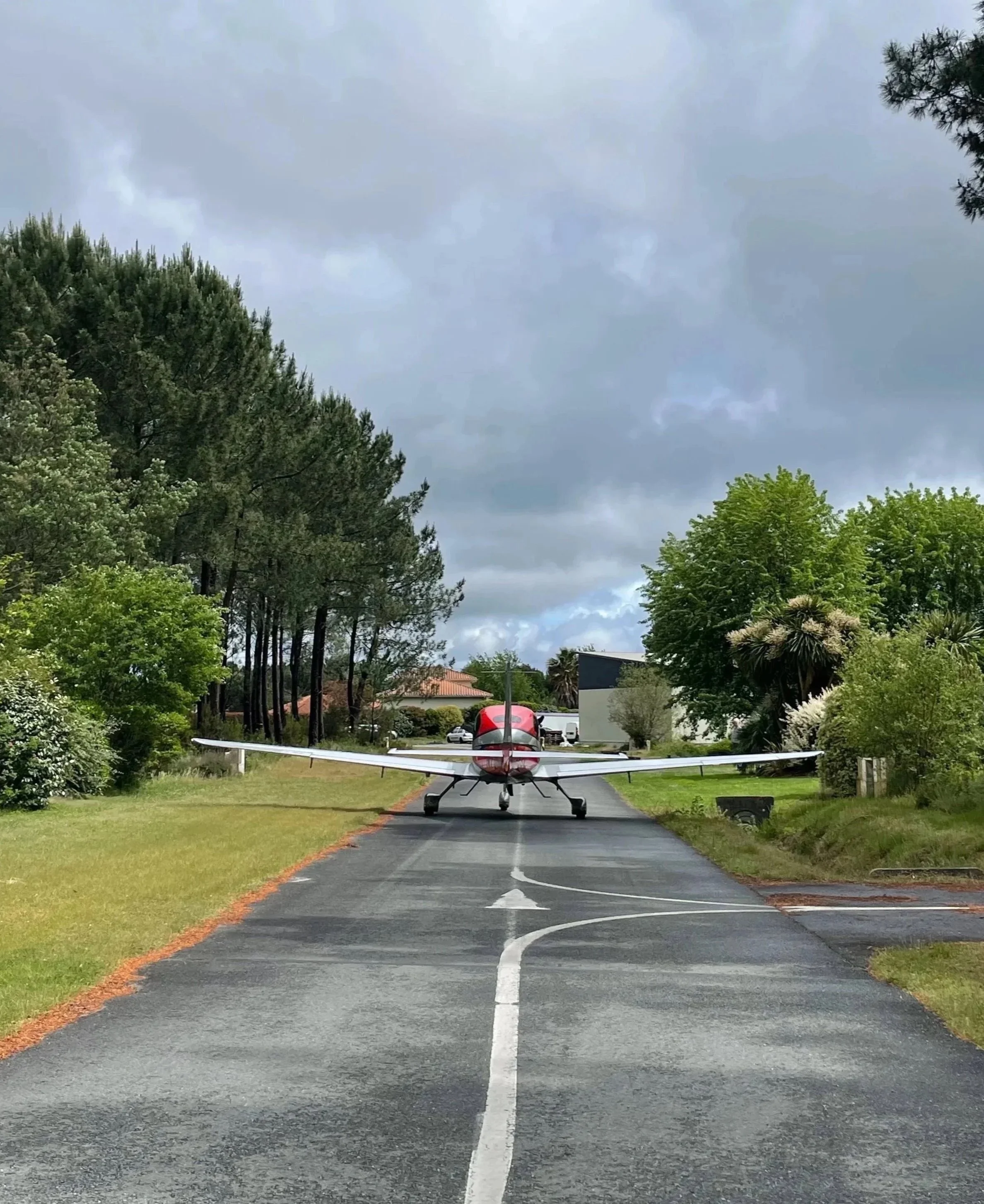 Un avion léger rouge et blanc circulant sur une un taxiway dans un village aéronautique privé, bordé d'arbres et de maisons, avec un ciel nuageux.