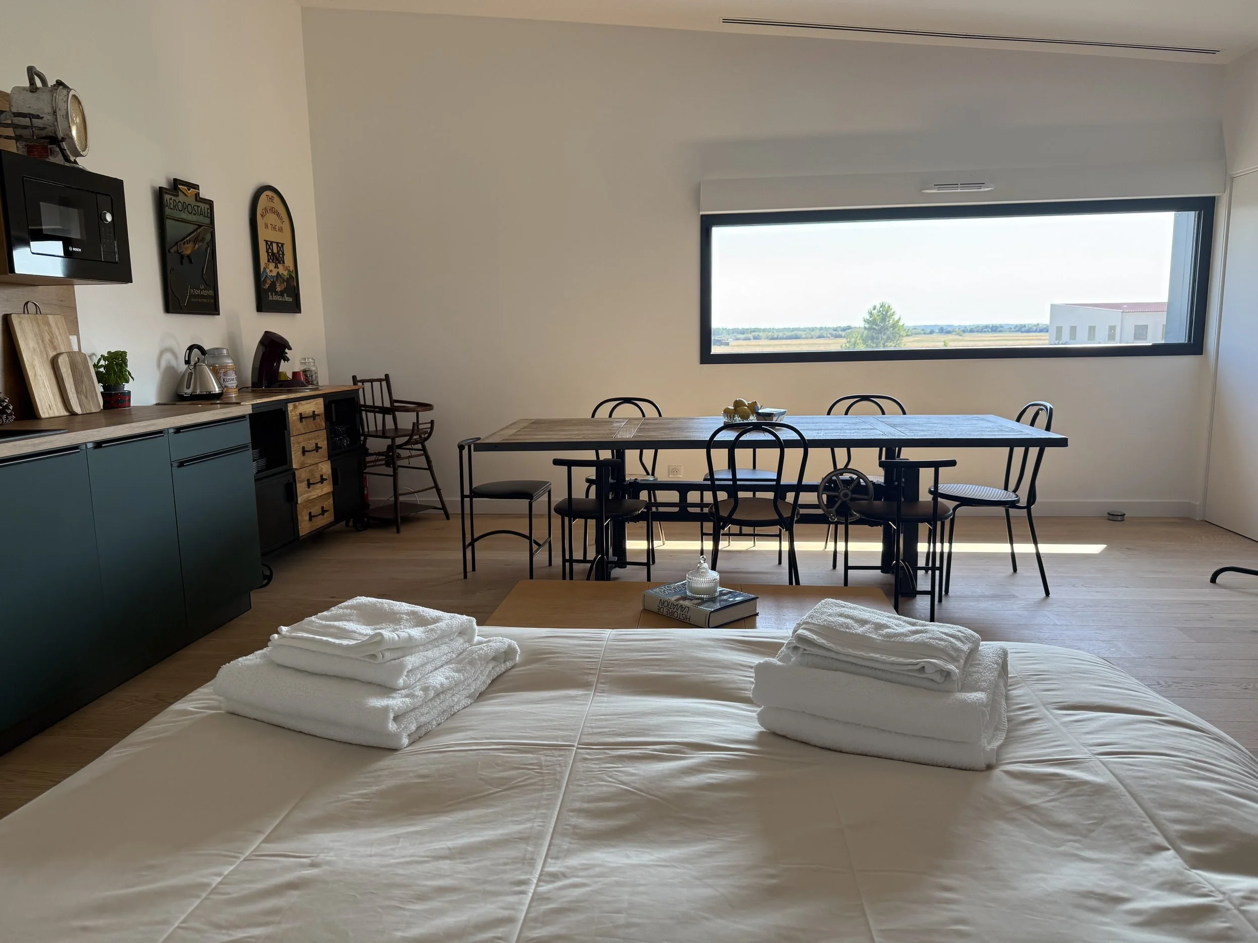 Chambre avec lit, serviettes pliées sur le lit, grande fenêtre laissant entrer la lumière, table à manger avec six chaises, cuisine en arrière-plan.