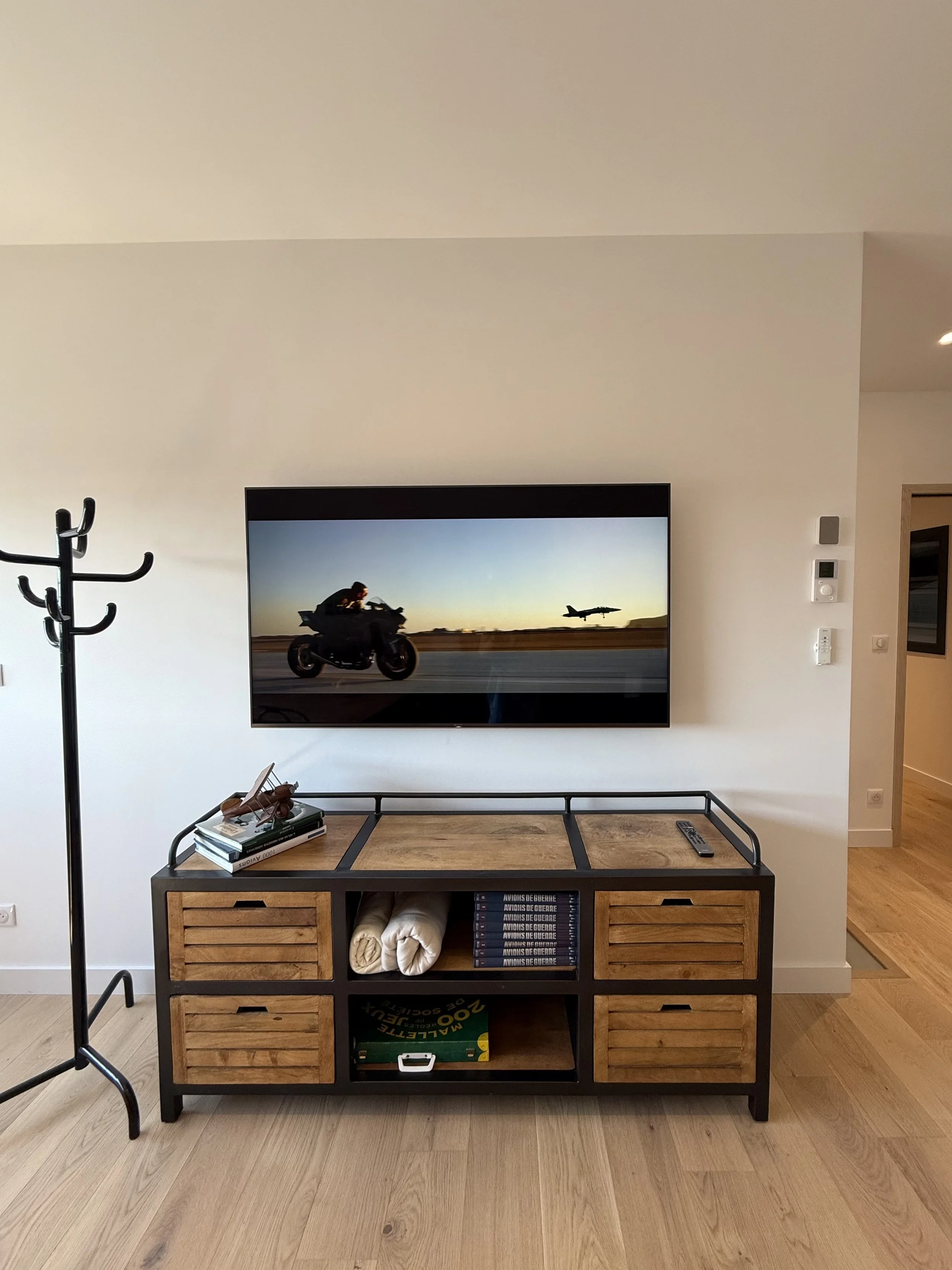 Salon avec une télévision murale, meuble en bois et métal, livres, une représentation d'un avion au sol et un avion en vol à l'écran, sur un mur blanc.
