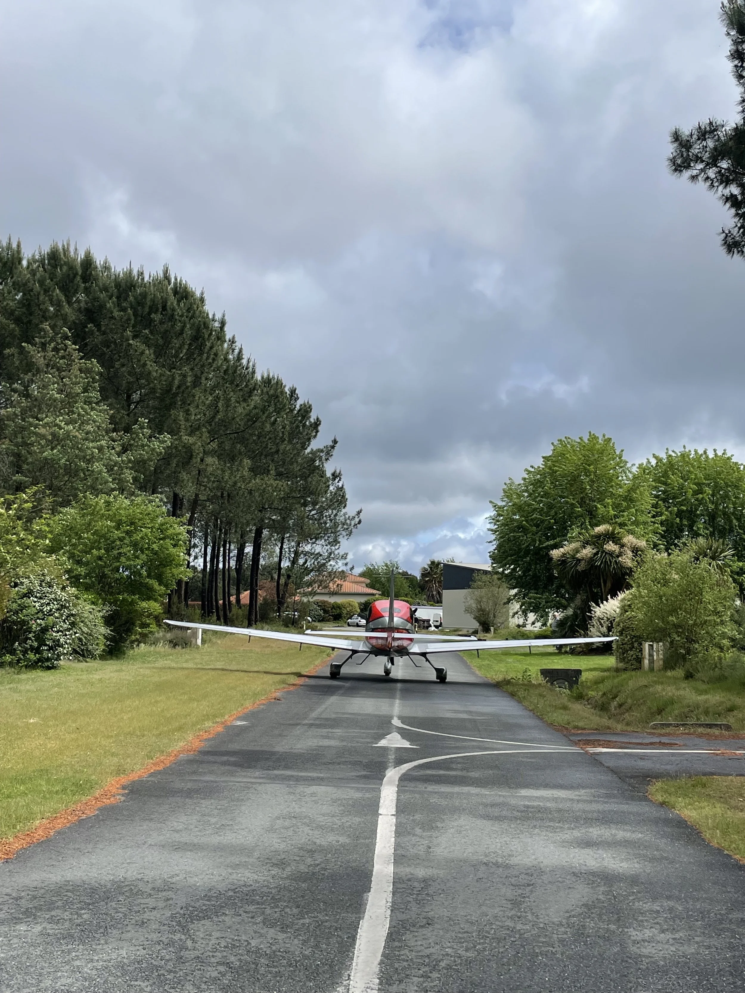 Un petit avion circule au milieu du village aéronautique de Biscarrosse, sur une route pavée, entouré d'arbres verts et de maisons, avec un ciel nuageux au-dessus.