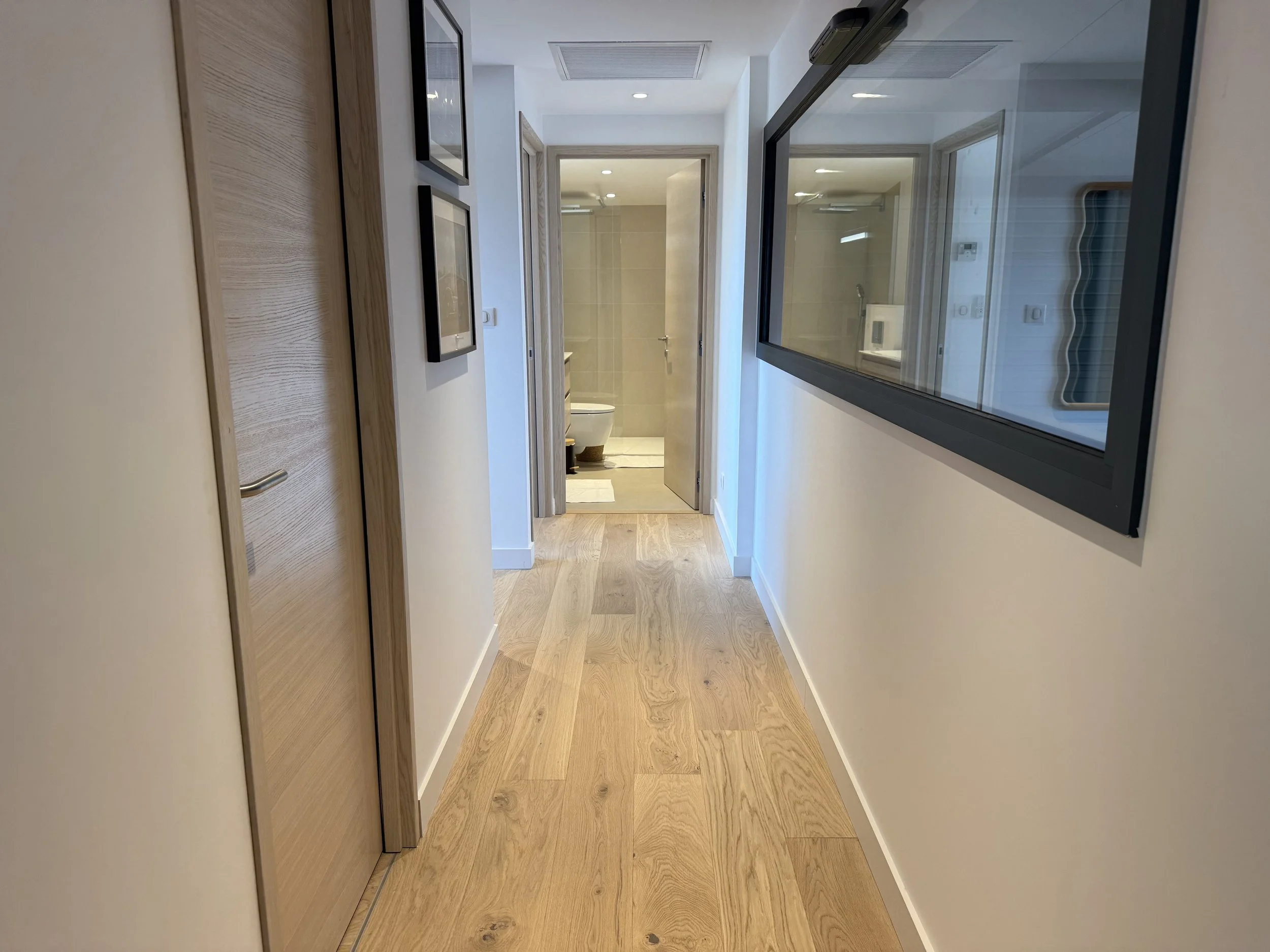 Couloir dans un appartement moderne avec portes en bois, mur blanc, et un grand miroir rectangulaire sur le mur de droite. Au bout du couloir, il y a une salle de bain avec des toilettes visibles.