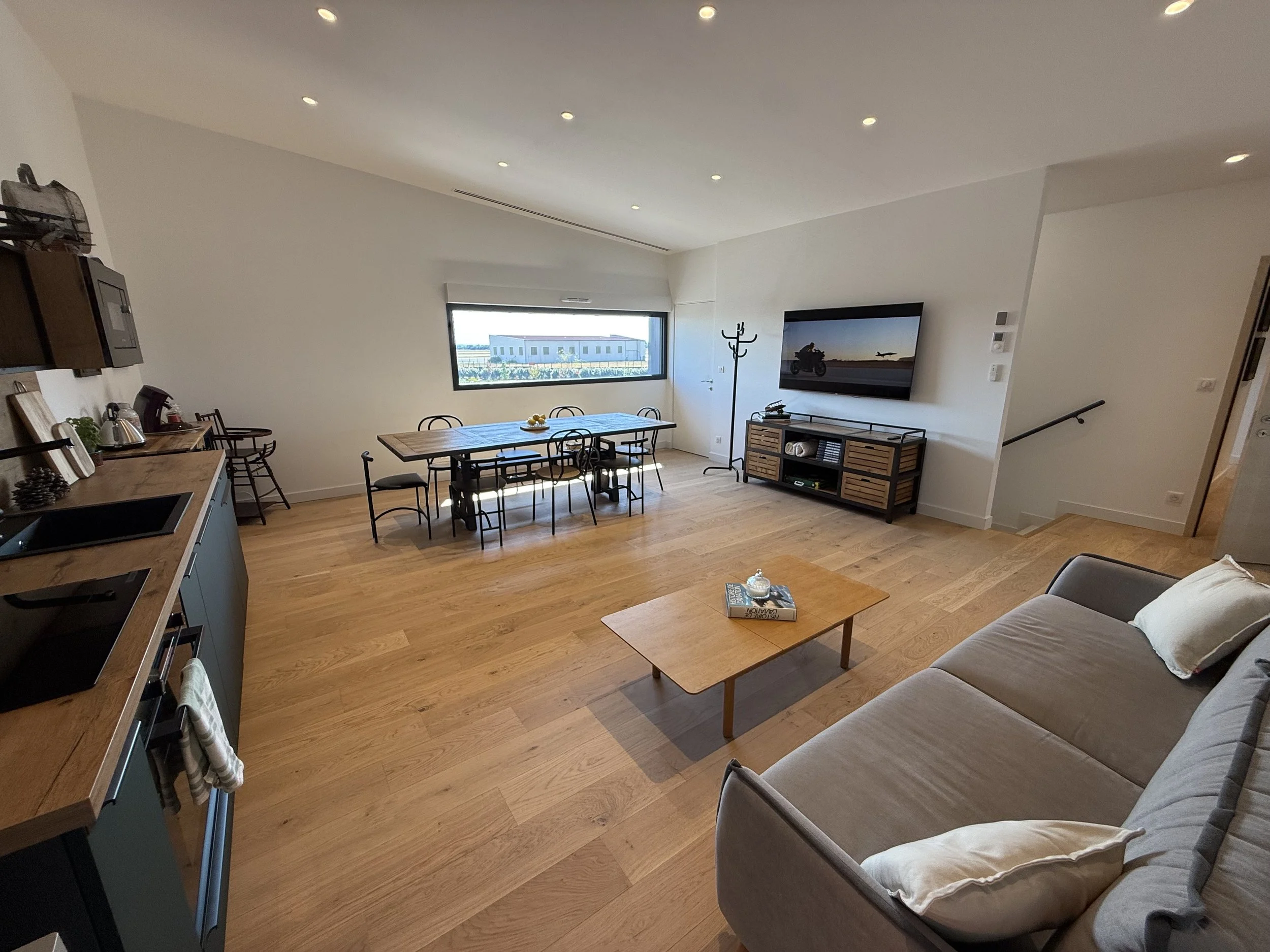 Salon intérieur moderne avec canapé gris, table basse en bois, télévision murale, table à manger en bois entourée de chaises, cuisine en arrière-plan, et grande fenêtre laissant entrer la lumière naturelle.