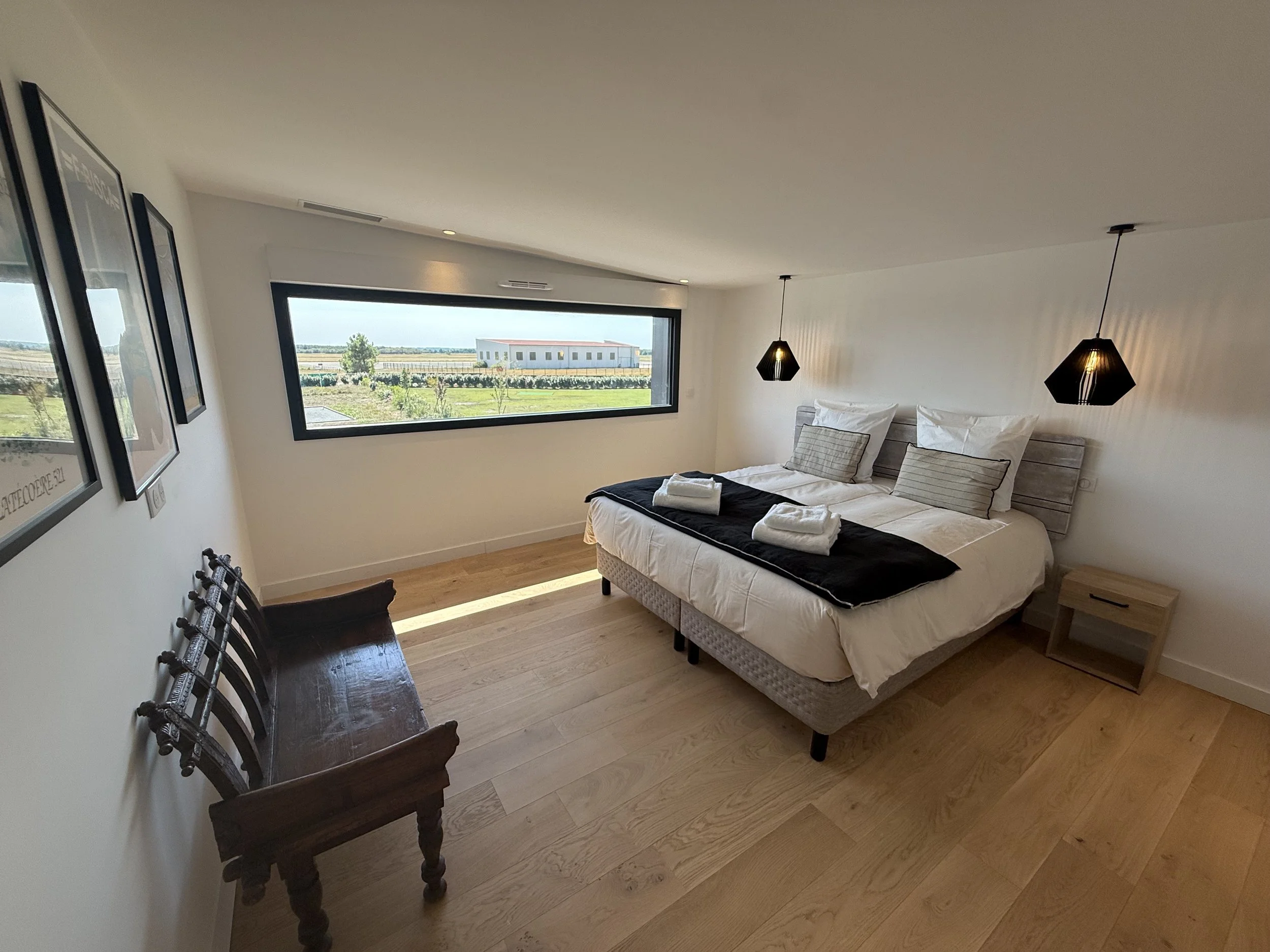 Chambre moderne avec lit double, deux lampes suspendues, grande fenêtre, et mobilier en bois, vue sur un paysage rural.