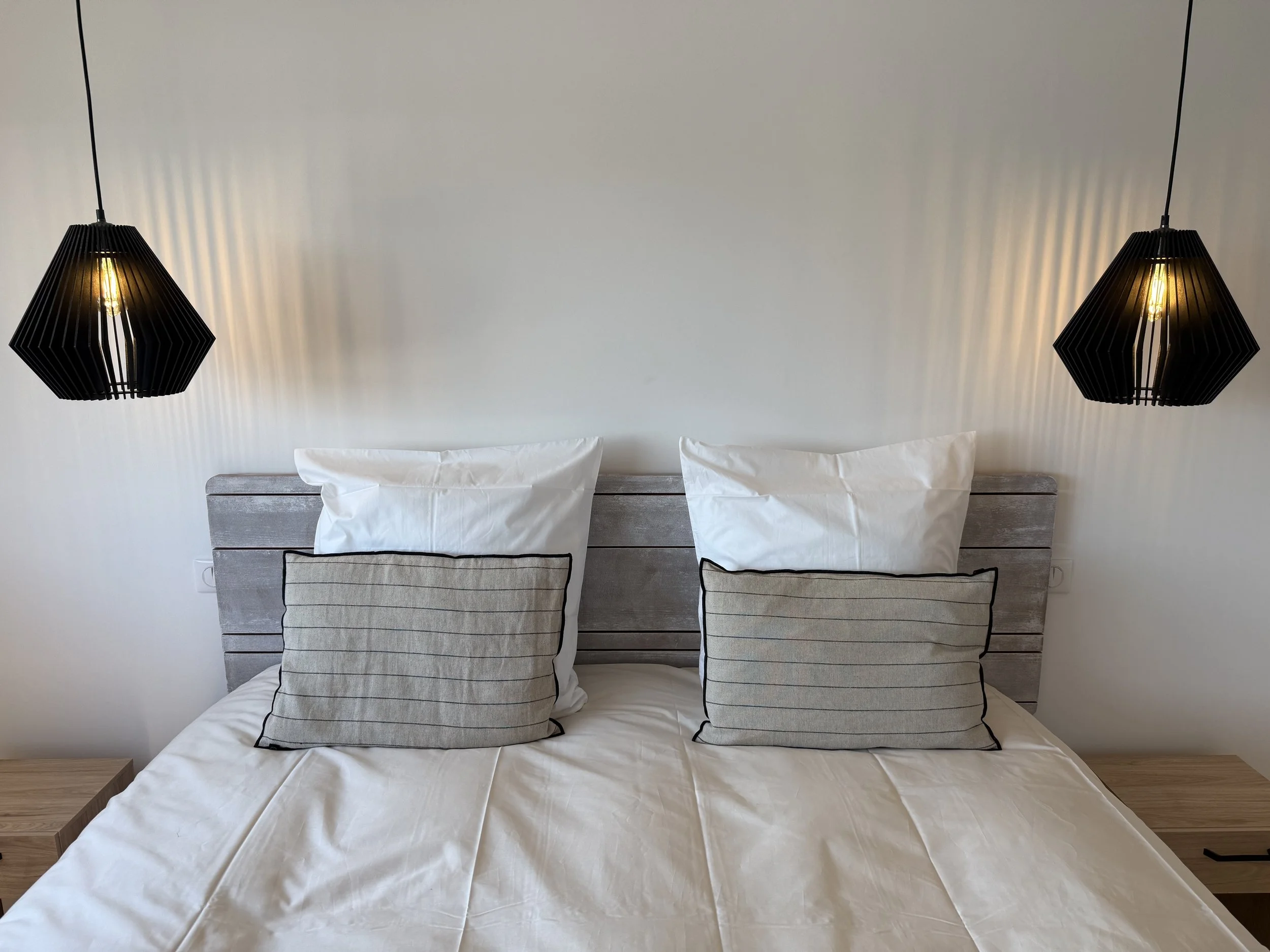 Lit de chambre avec deux oreillers blancs et deux coussins gris à rayures, à côté de deux lampes suspendues noires.