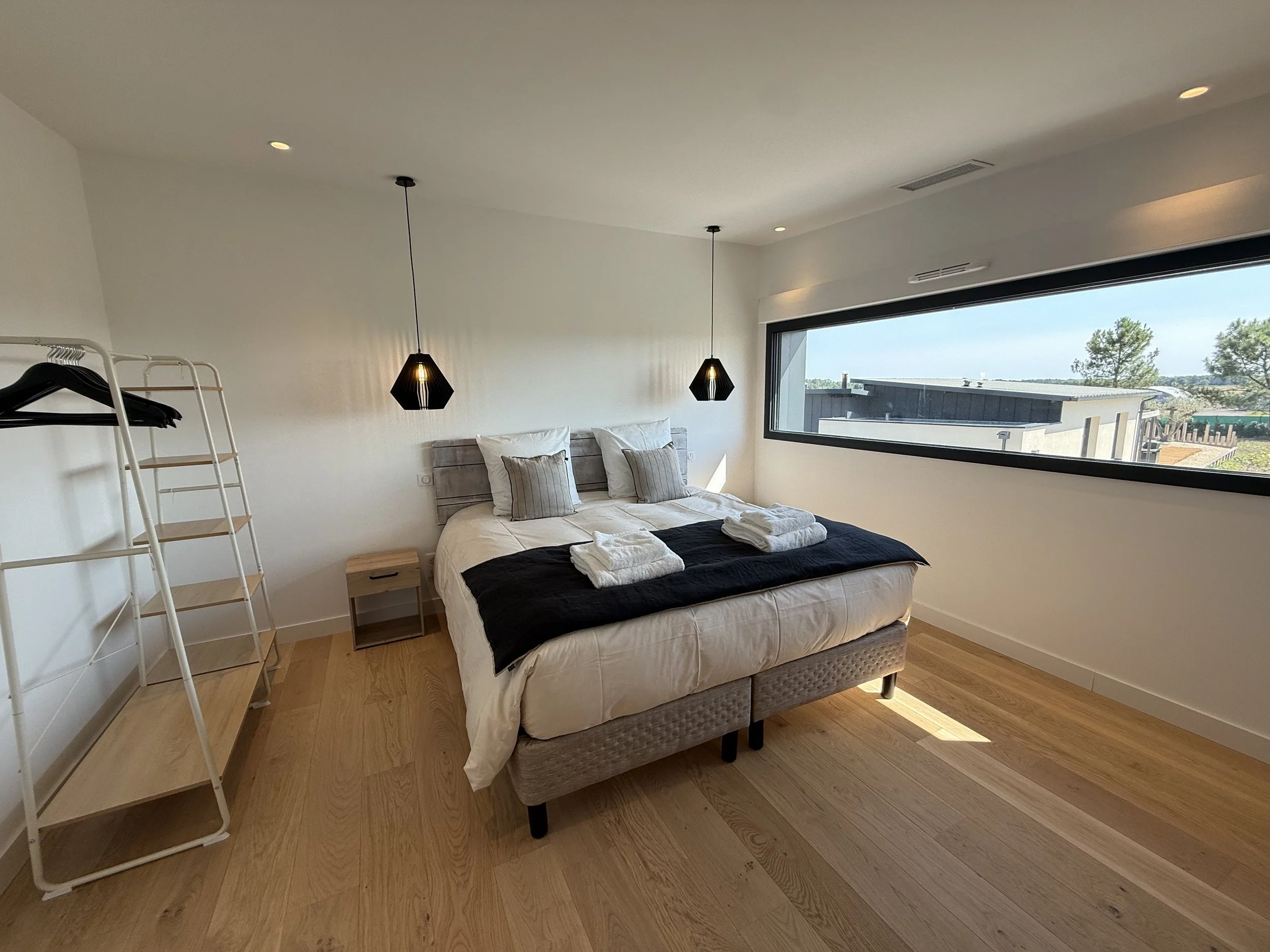 Chambre moderne avec lit double, deux lampes suspendues, fenêtre longue et vue sur des arbres, parquet en bois clair, armoire blanche et petite table de nuit.