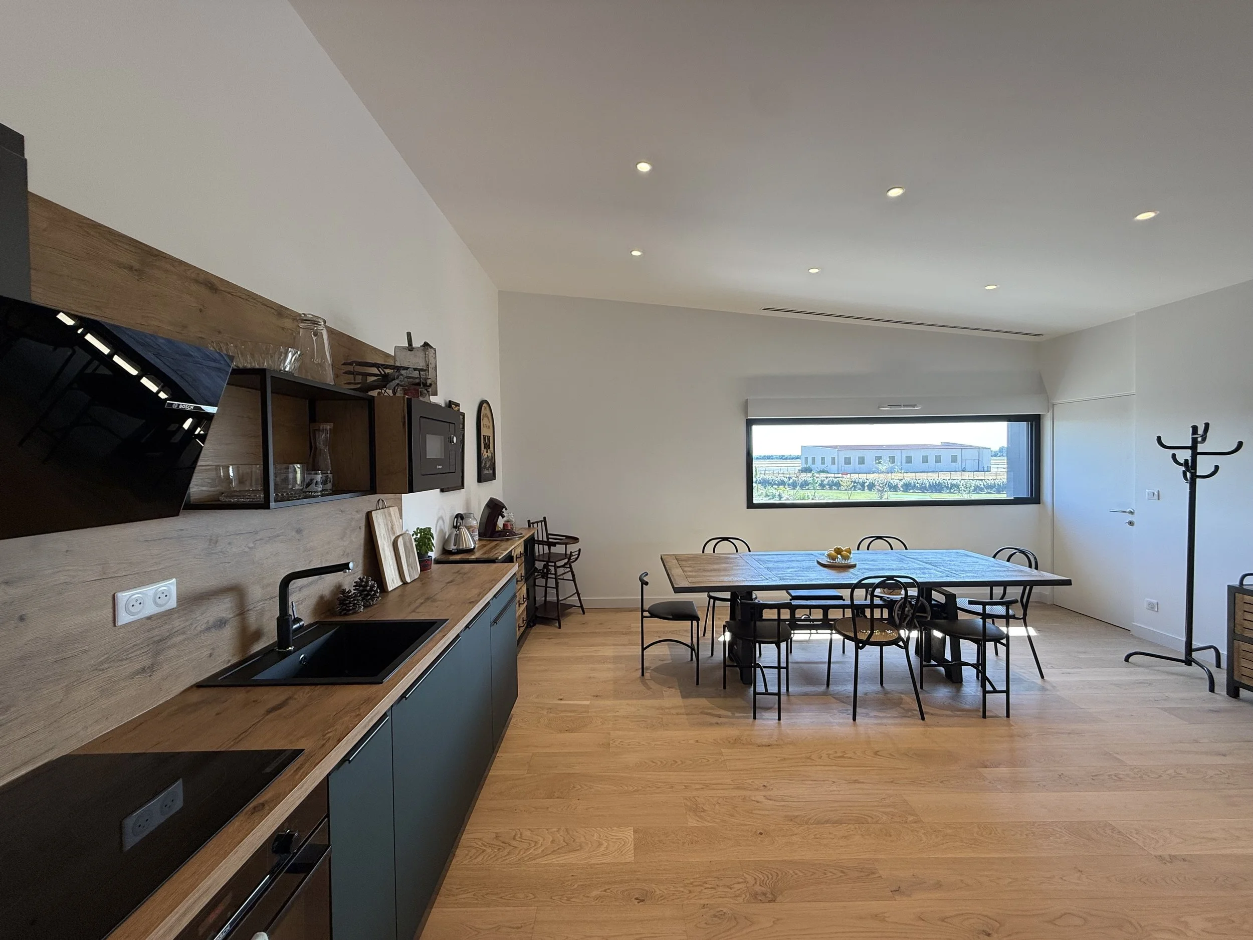 Cuisine et salle à manger modernes avec plan de travail en bois, meubles noirs, grande fenêtre offrant une vue sur un bâtiment moderne, table en bois entourée de chaises en métal, porte blanche et porte-manteau noir.