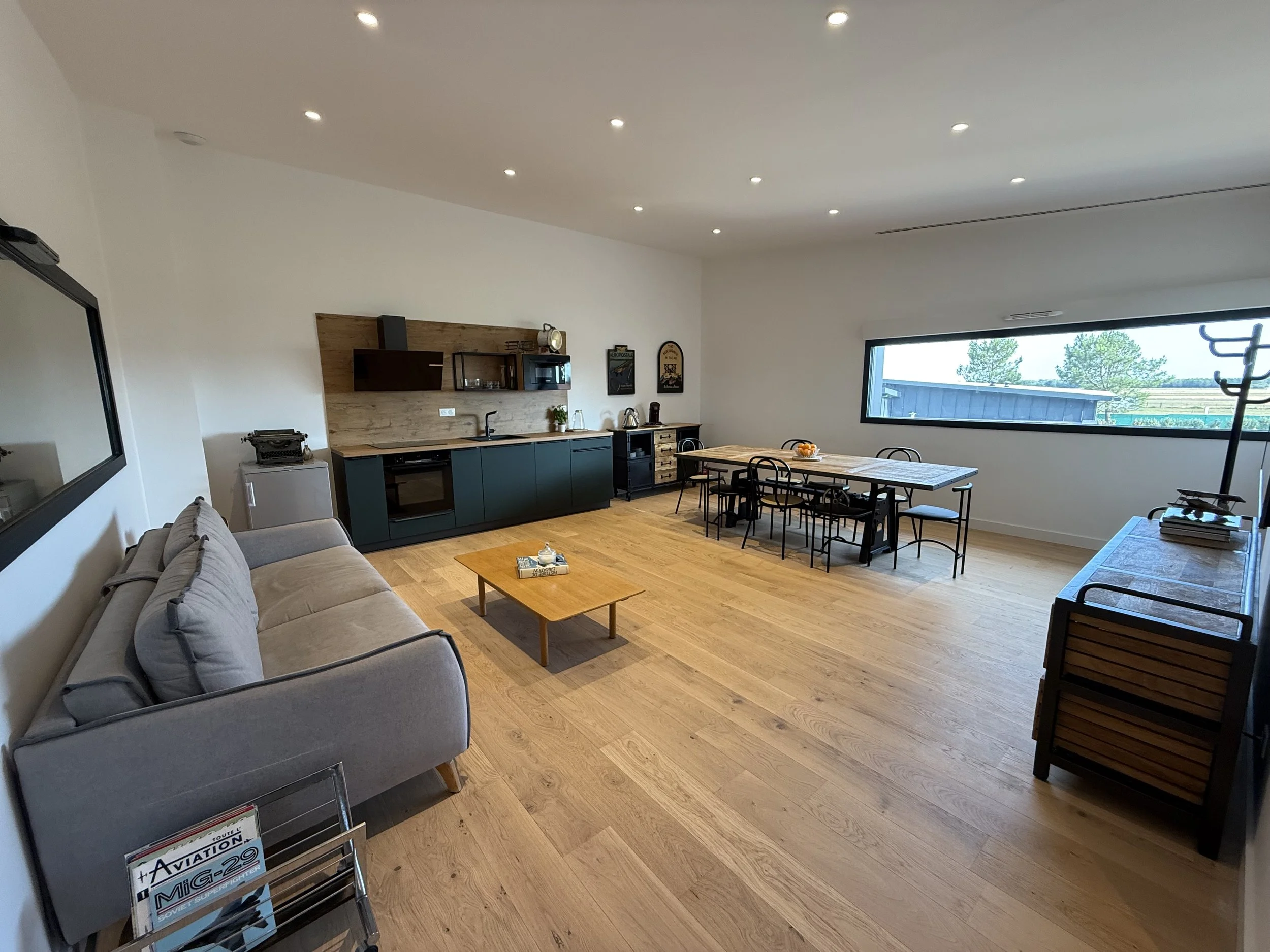 Salle de séjour moderne avec canapé gris, table basse, cuisine ouverte avec armoires vertes et plan de travail en bois, grand espace avec table à manger et chaises, grande fenêtre offrant une vue sur la nature.