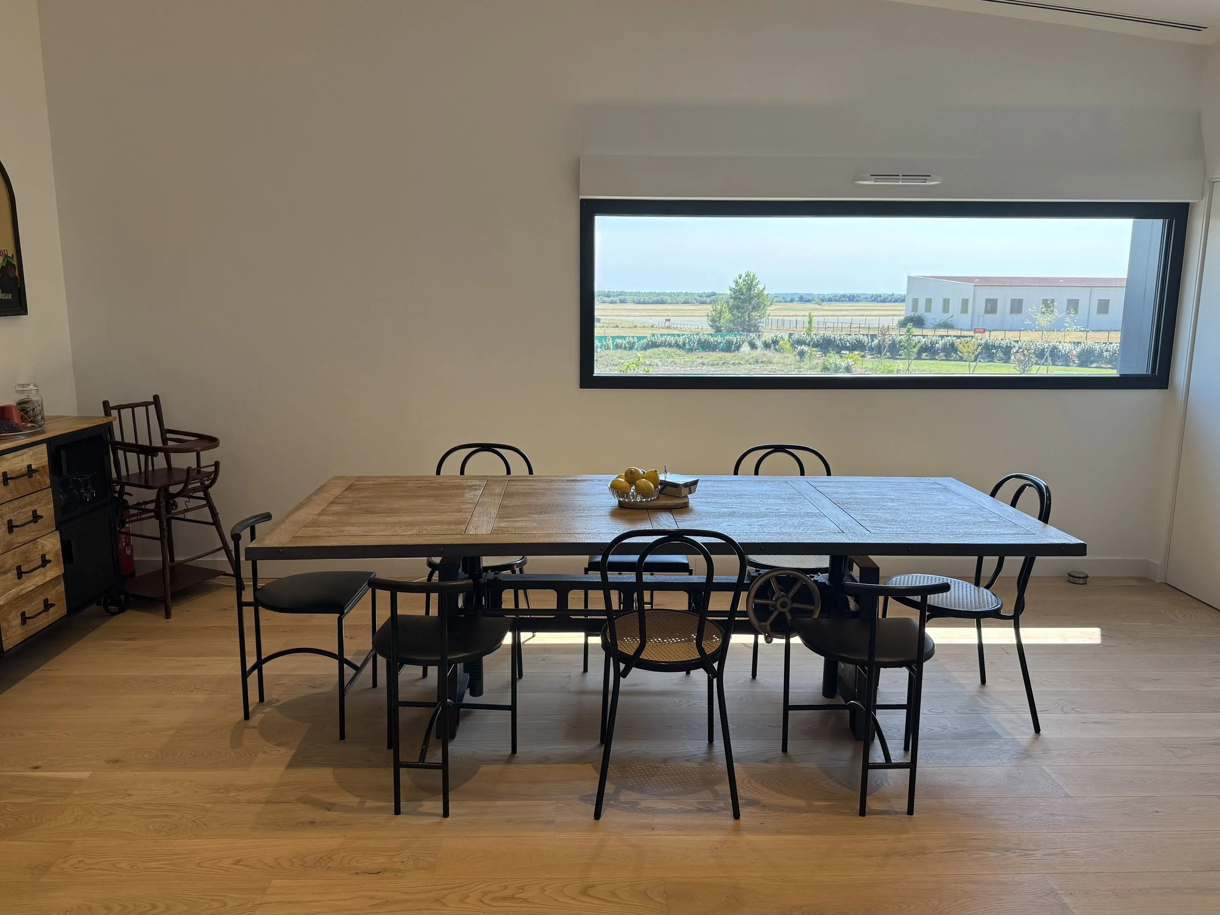 Salle à manger avec une grande table en bois entourée de six chaises noires et une grande fenêtre offrant une vue sur un paysage agricole.