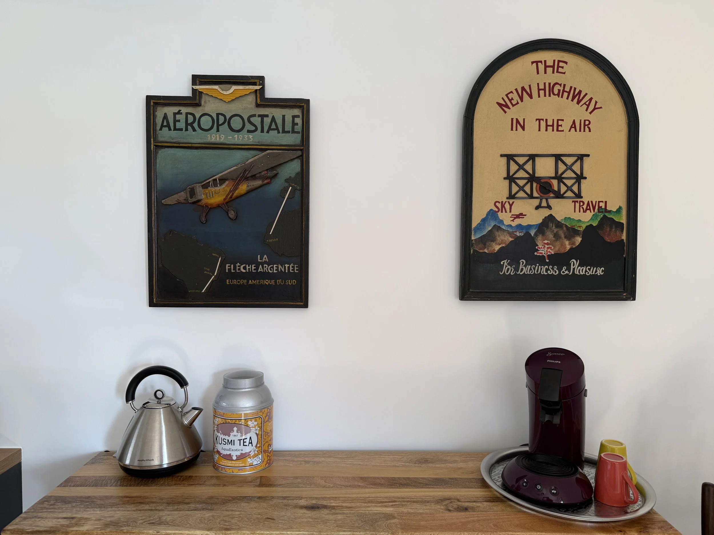 Une cheminée électrique, une théière en acier inoxydable, une boîte de thé KUSMI TEA, une machine à café rouge avec une soucoupe, deux tasses rouges et jaunes sur une table en bois face à un mur blanc avec deux affiches vintage.