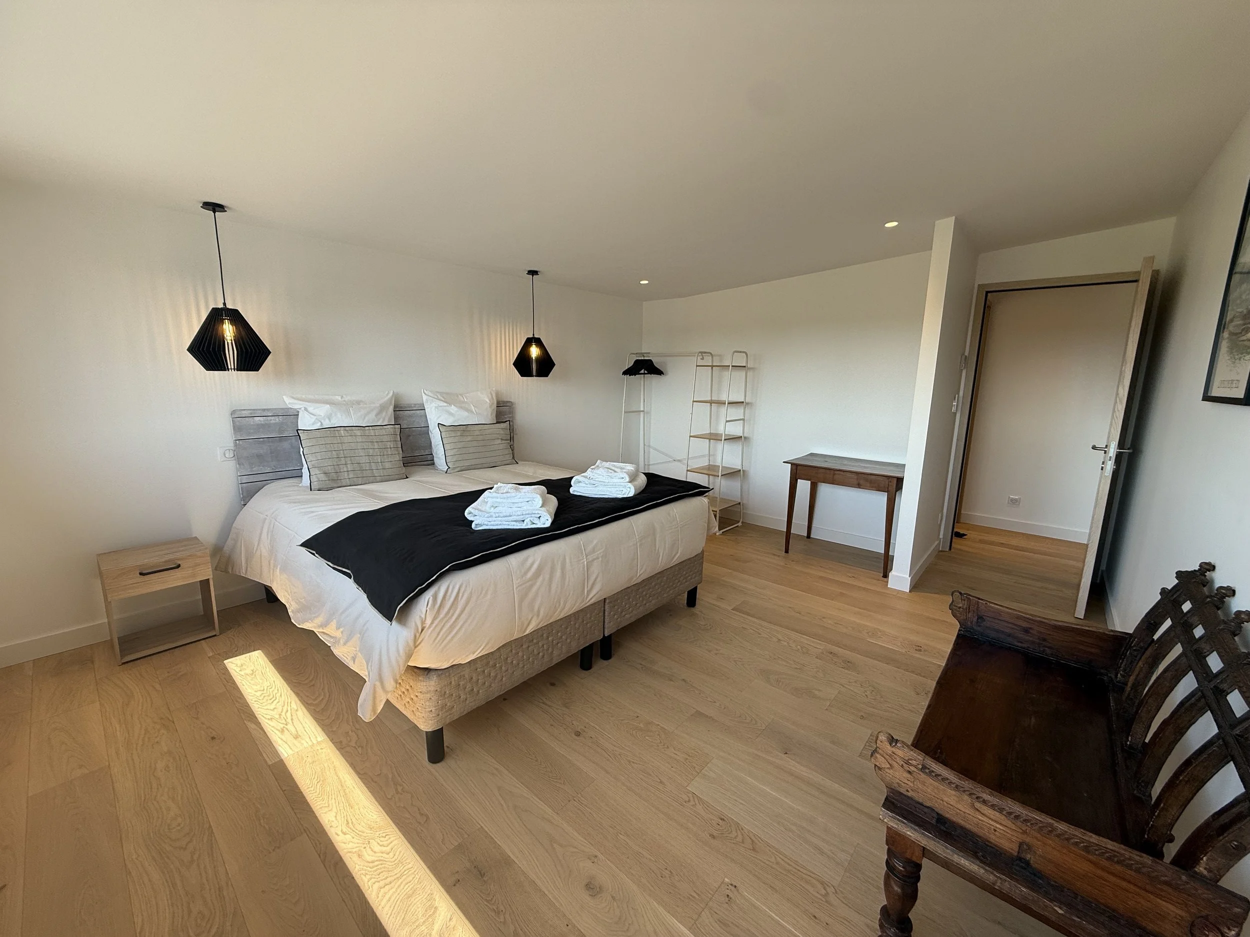 Chambre de chambre moderne avec lit double, draps blancs, coussins, lampes suspendues noires, table de chevet en bois, étagère blanche, petite table en bois, porte entrouverte, fauteuil en bois ancienne.