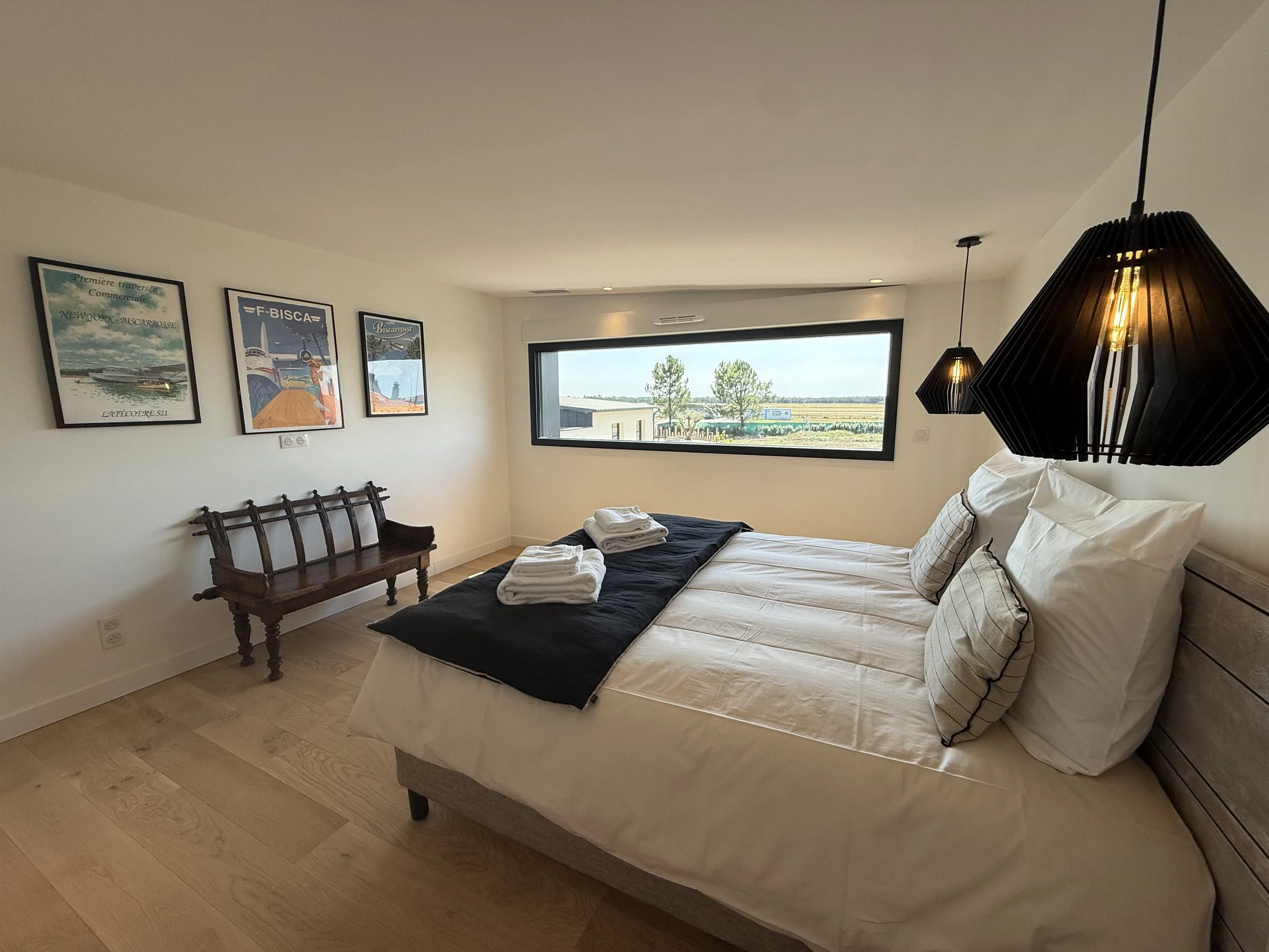 Chambre moderne avec lit double, lampes suspendues noires, tableau vintage, fenêtre offrant vue rurale, serviettes pliées sur le lit.