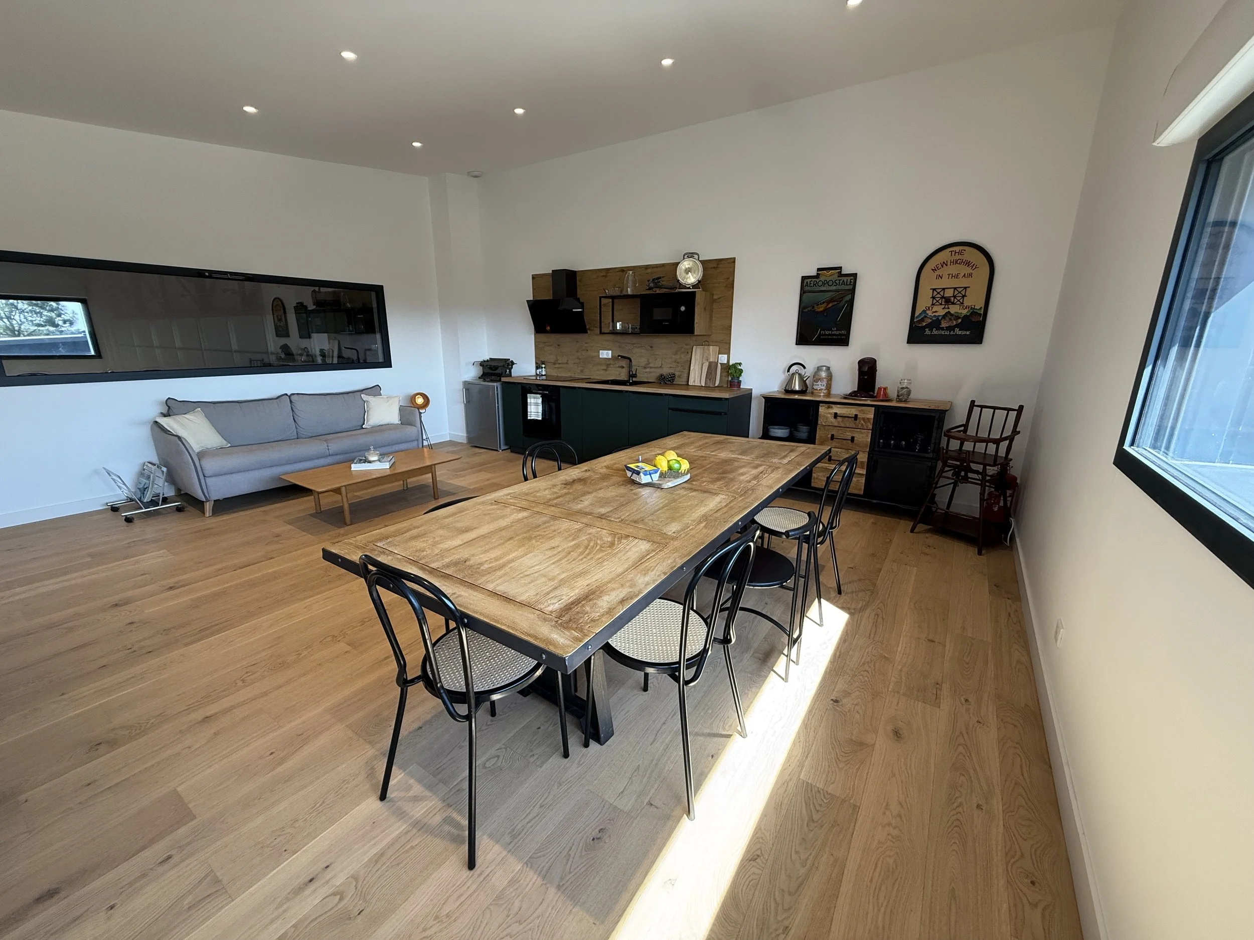 Salle à manger avec une grande table en bois, six chaises noires, un canapé gris avec deux coussins blancs, une petite table en bas à gauche, une cuisine équipée derrière avec des armoires vertes, des hôtes en bois, un micro-ondes, quelques affiches