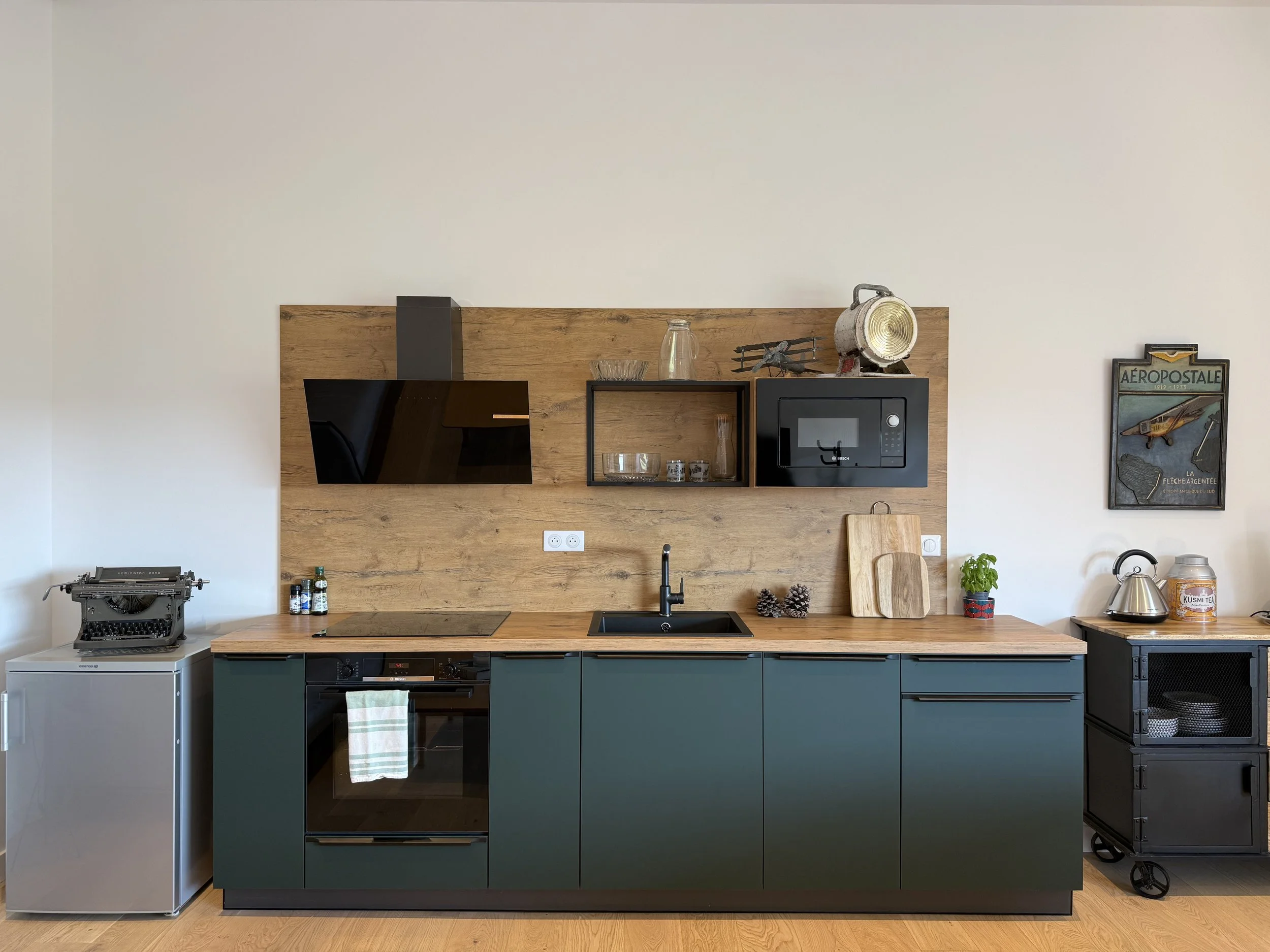 Cuisine moderne avec plan de travail en bois, appareils électroménagers noirs, micro-ondes, évier, porte-texte vintage sur le mur, petits accessoires de cuisine et une petite plante.