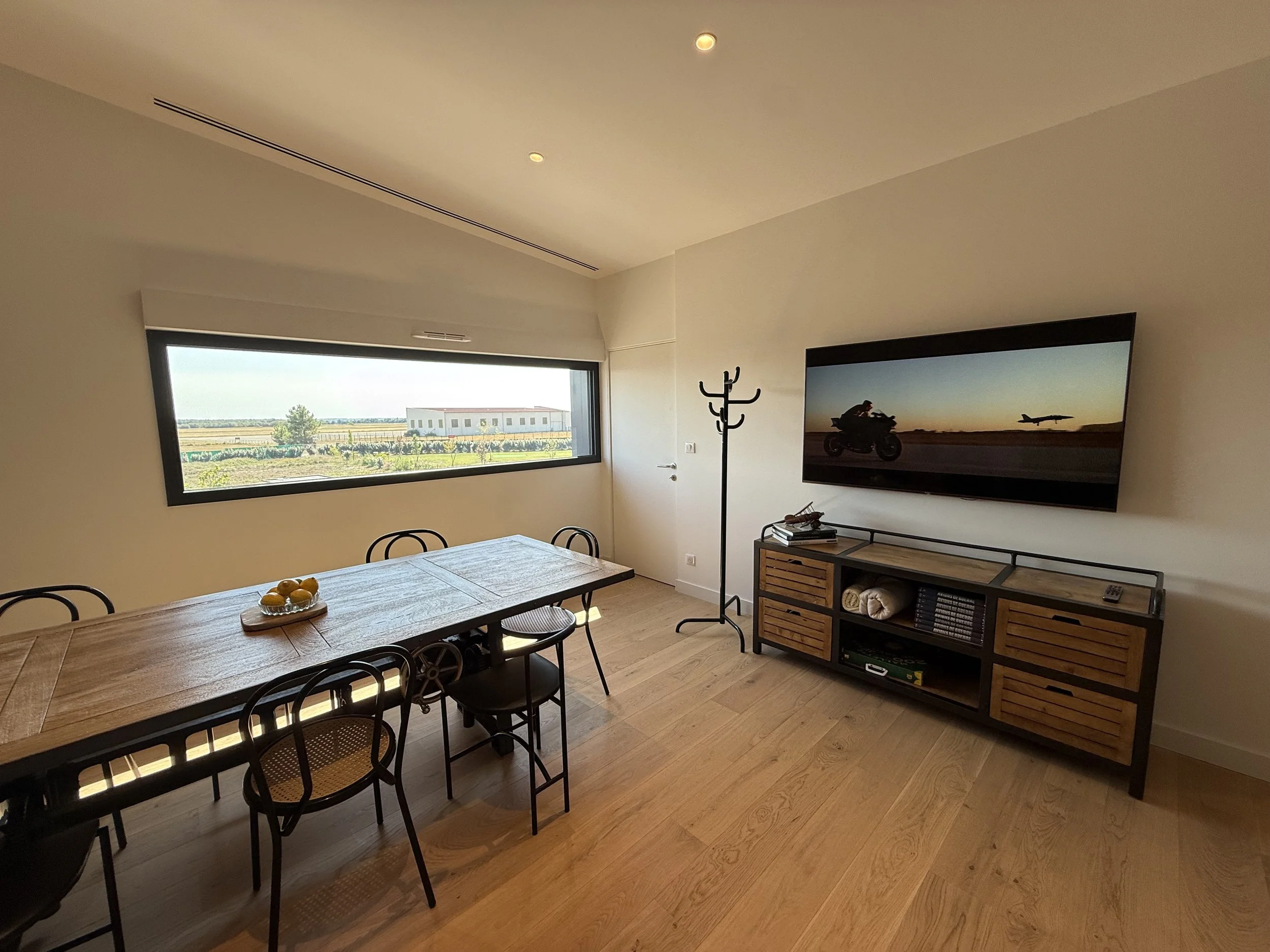Salle à manger avec table en bois, chaises noires, grande fenêtre avec vue sur un terrain et bâtiments, télévision murale affichant une scène de moto et avion, meuble de rangement en bois et métal, porte, porte-manteau et sol en bois clair.
