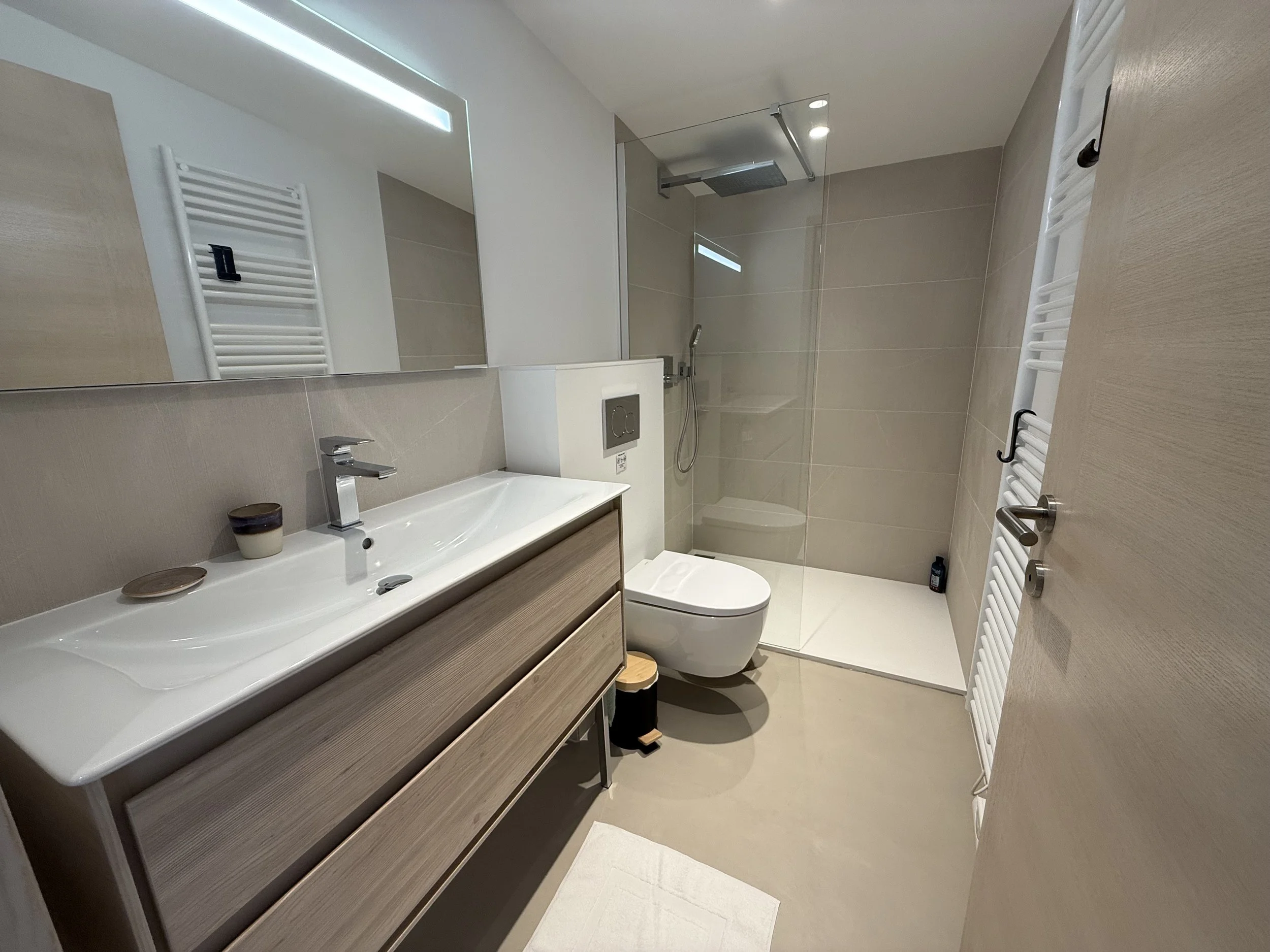 Salle de bain moderne avec un lavabo en bois clair, miroir, toilette suspendue, douche à l'italienne avec paroi en verre et douchette à pluie, radiateur sèche-serviettes blanc, et accessoires en bois et en céramique.