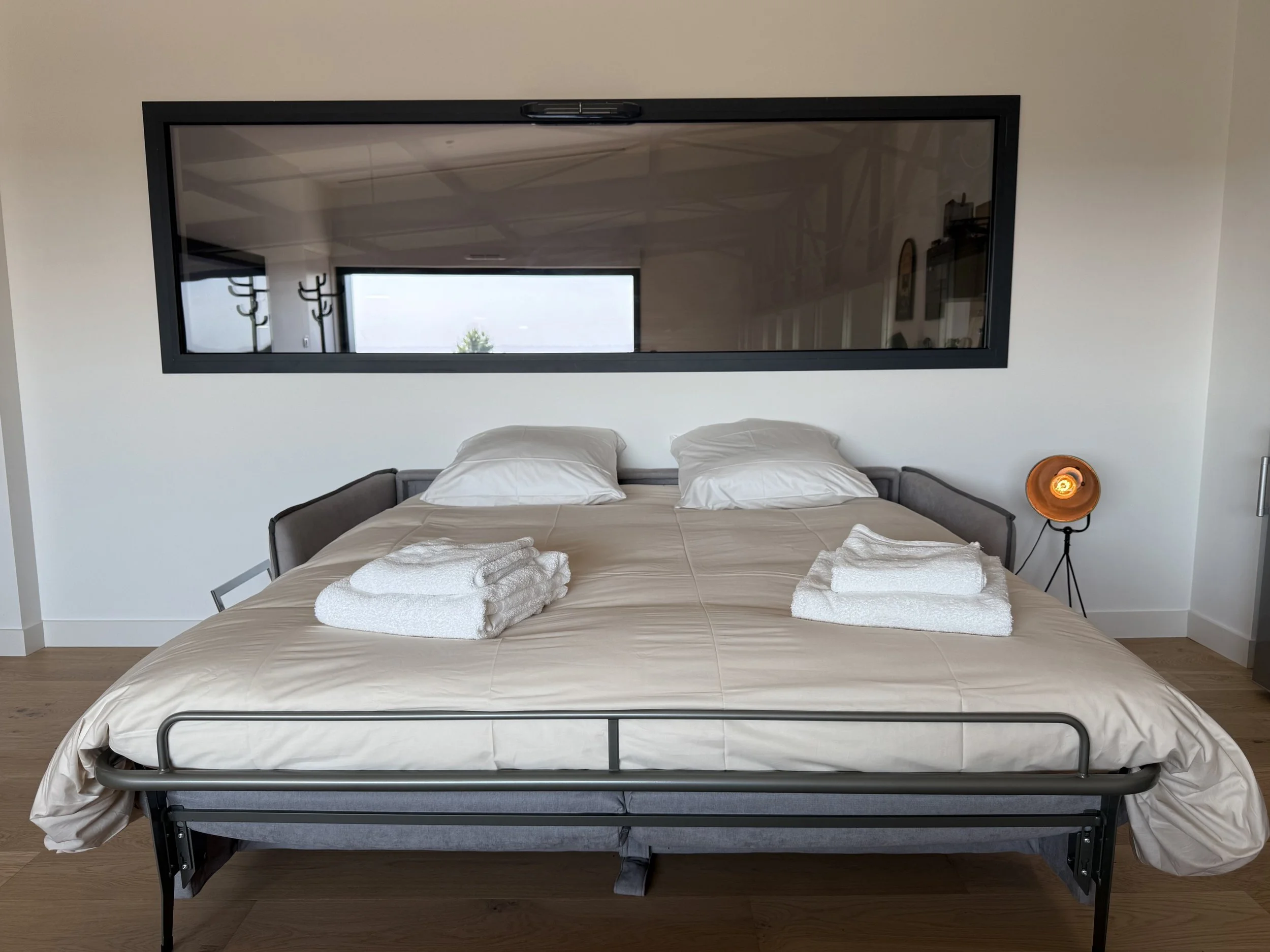 Lit double avec deux coussins, serviettes blanches pliées, miroir noir rectangulaire au mur, lampe de chevet orange à droite, parquet en bois, mur blanc.