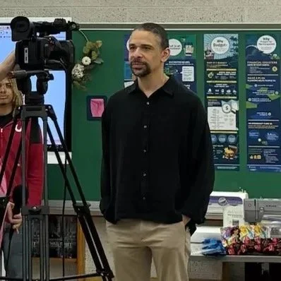 A man in a black shirt and beige pants being recorded on camera in a classroom with a green bulletin board in the background. Wilson Below Jr. Project Elevate 360 and Elevate 302.