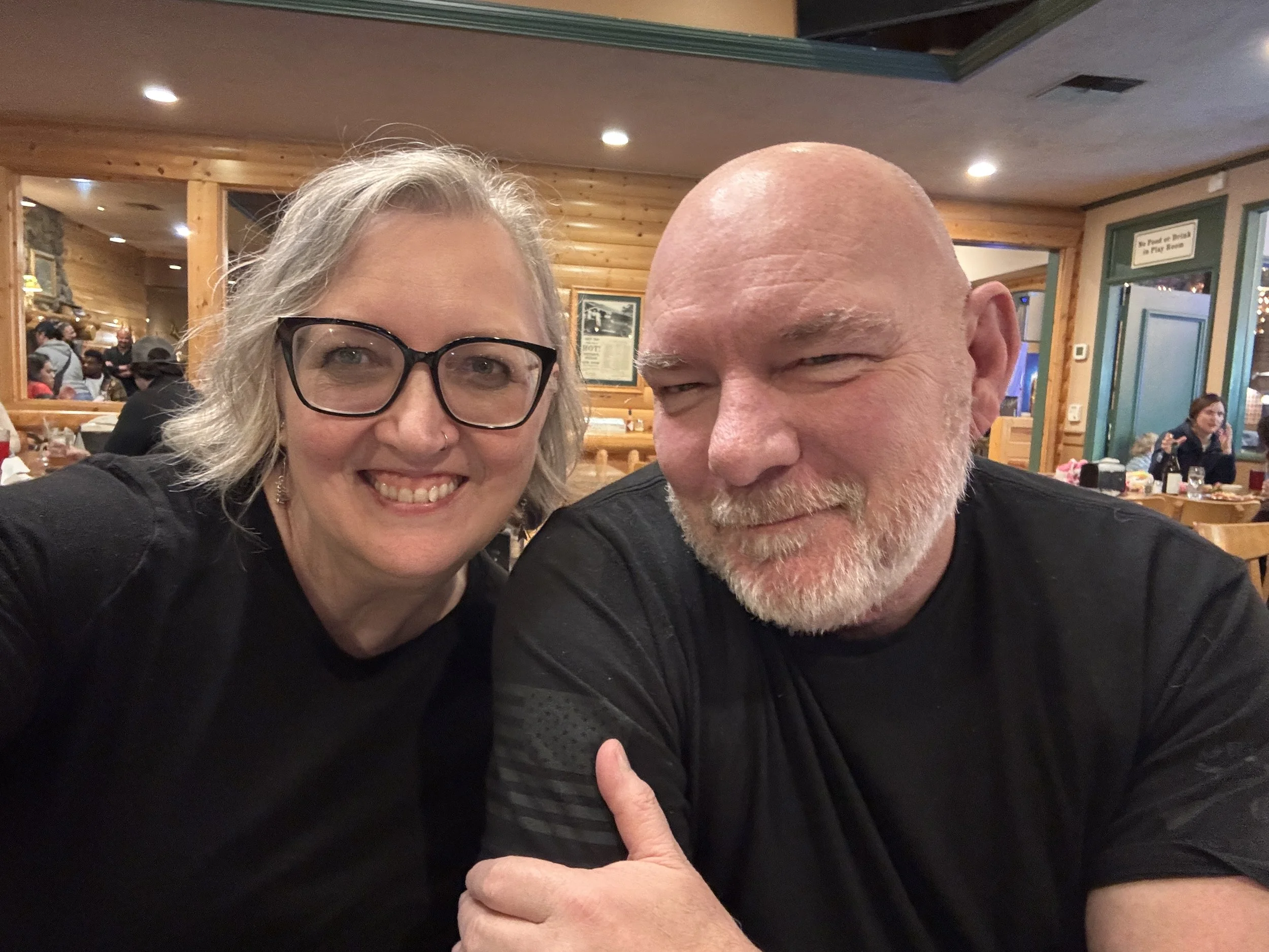 Sheila and Joe Morgan, husband and wife, both wearing black shirts and seated in a restaurant. Sheila owns Paint With Friends, a Eugene mobile paint and sip party service.