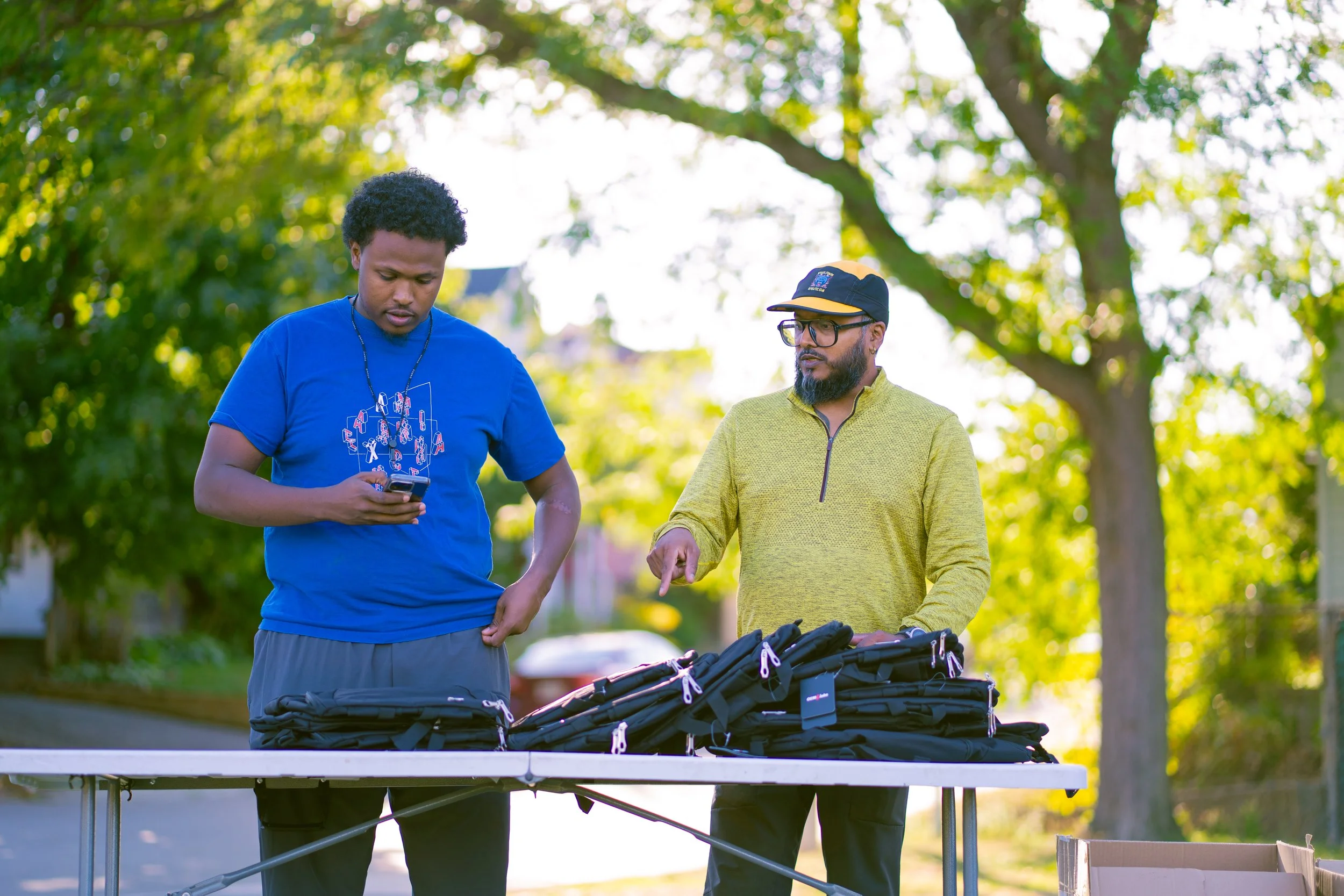 Two men are standing at a table outdoors, surrounded by trees; one is looking at his phone, and the other is pointing at a row of black backpacks on the table.