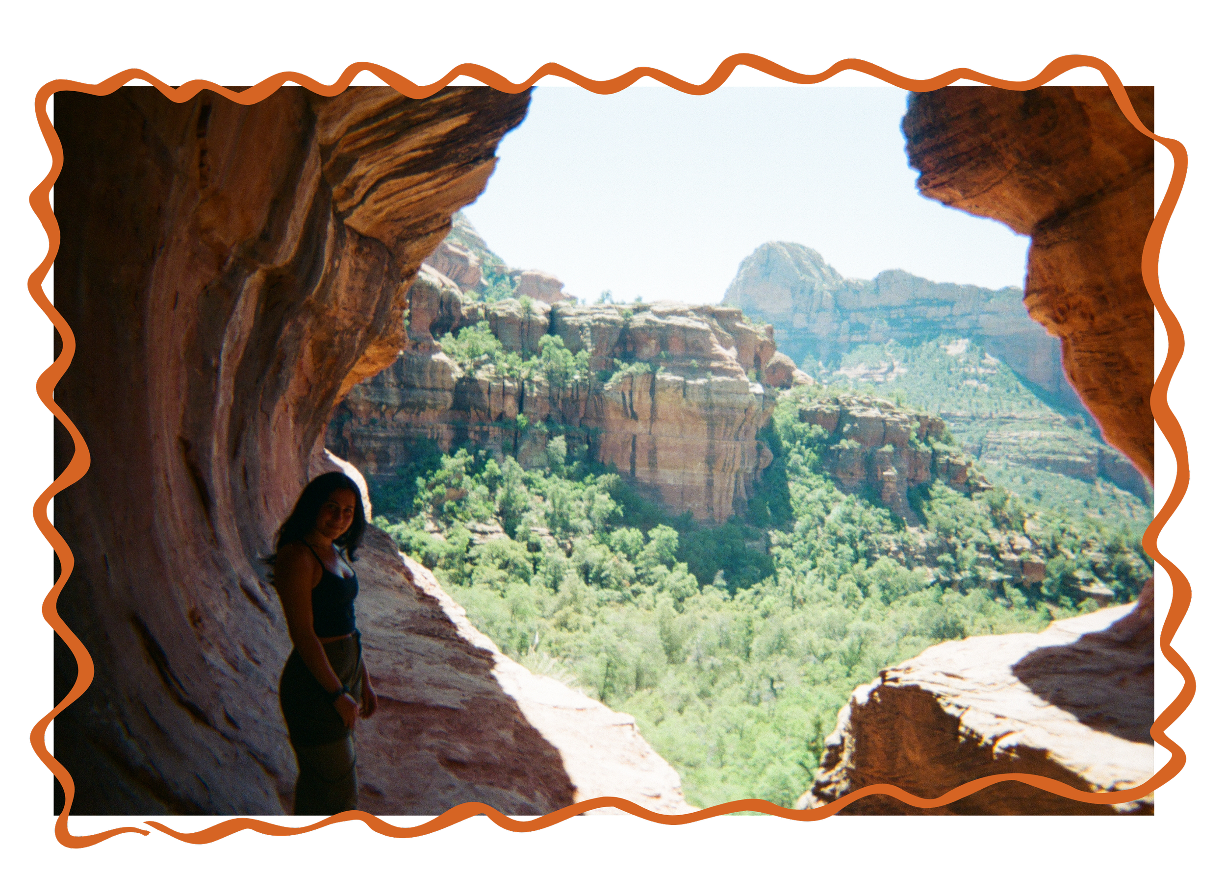Person standing inside a rock formation overlooking a canyon with red cliffs and green vegetation.