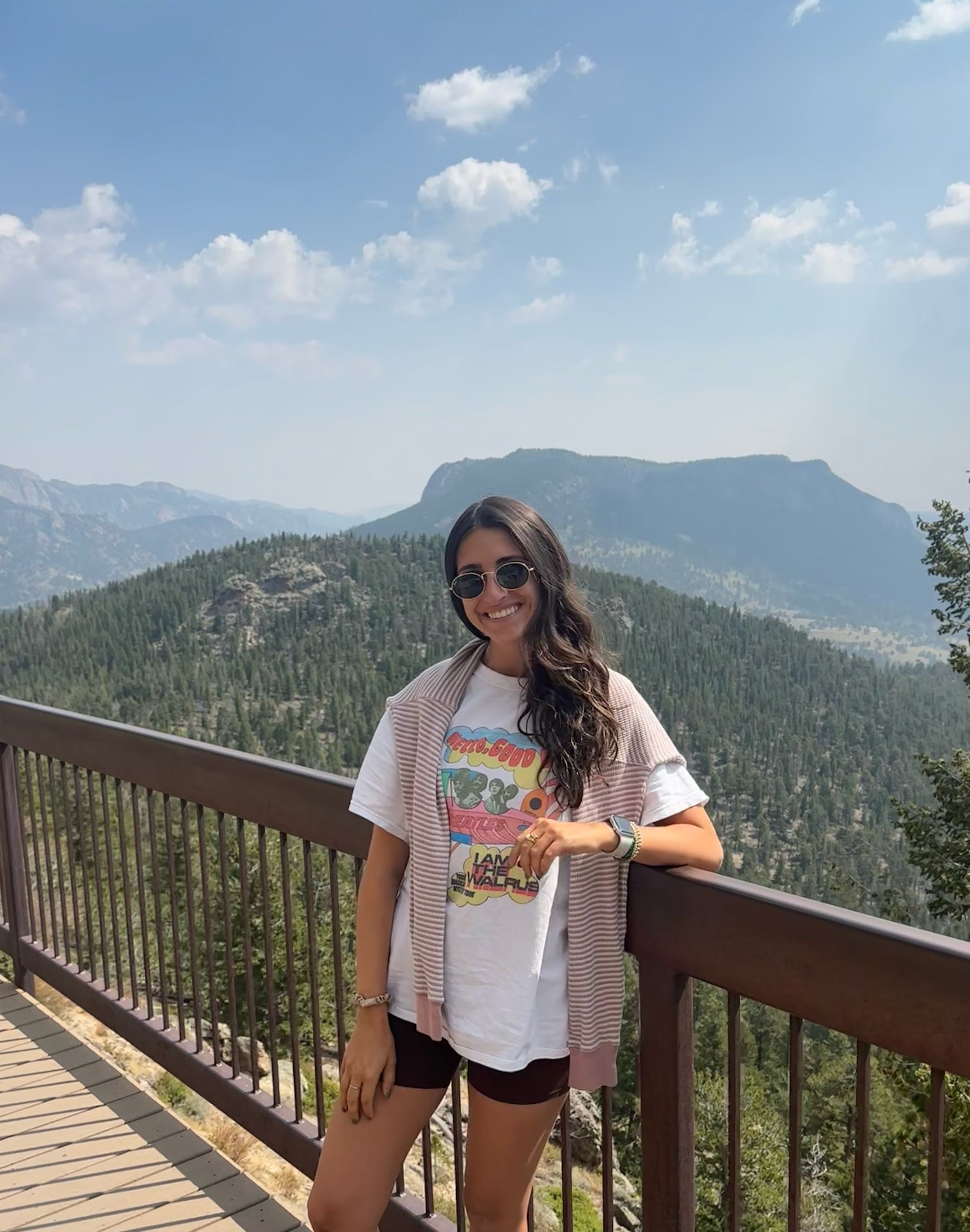 Woman smiling on a balcony overlooking a mountain landscape with trees, cliffs, and a partly cloudy sky.