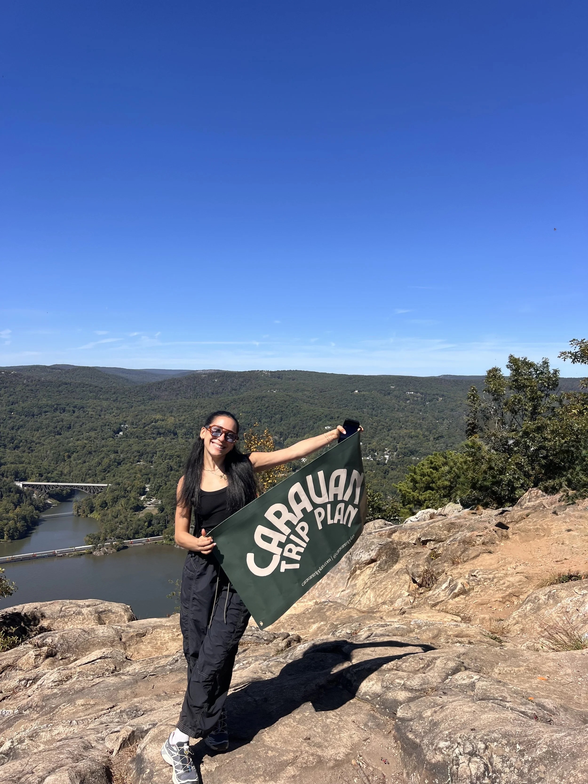 A woman standing on a rocky overlook holding a green flag that says "CARAUM TRIP PLAN" with a scenic view of a river, trees, and distant hills under a clear blue sky.