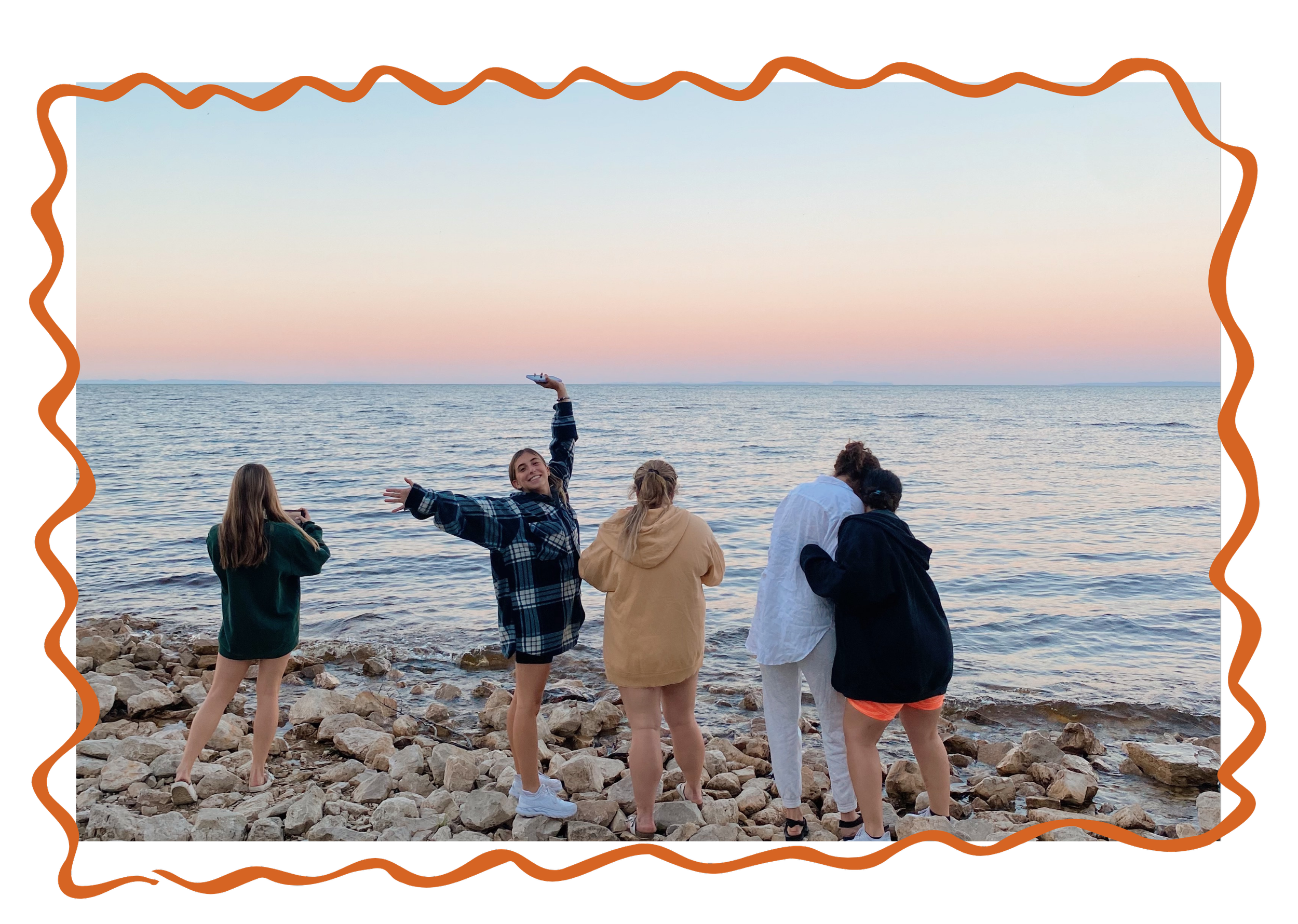 Group of five friends on a rocky beach taking photos and enjoying sunset over the water.