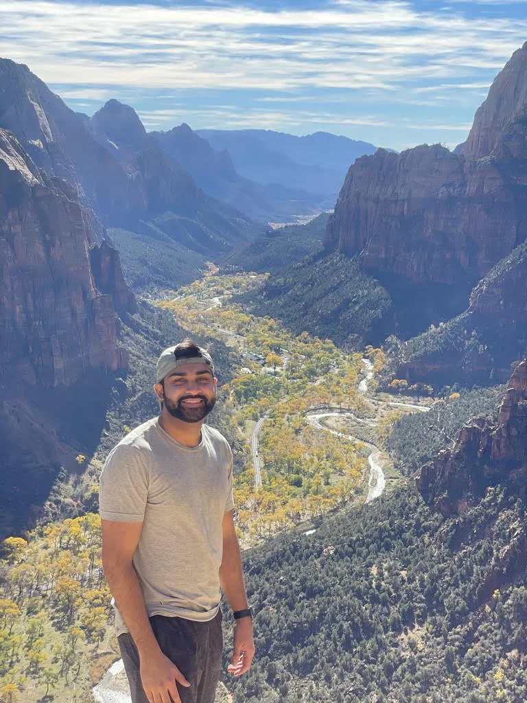 A man with a beard wearing a gray shirt and cap smiling at the camera, standing on a mountain overlook with a view of a valley, winding road, and tall cliffs in the background.