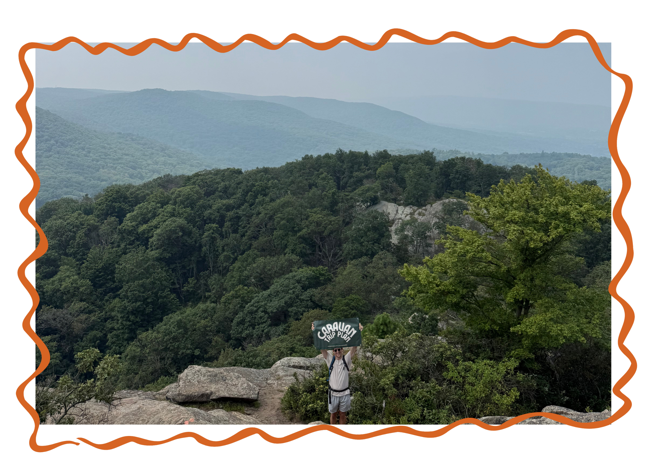 A person standing on a rocky trail at a mountain overlook, holding a sign that says 'Caravan.' They are surrounded by green trees and misty mountain ranges in the background.