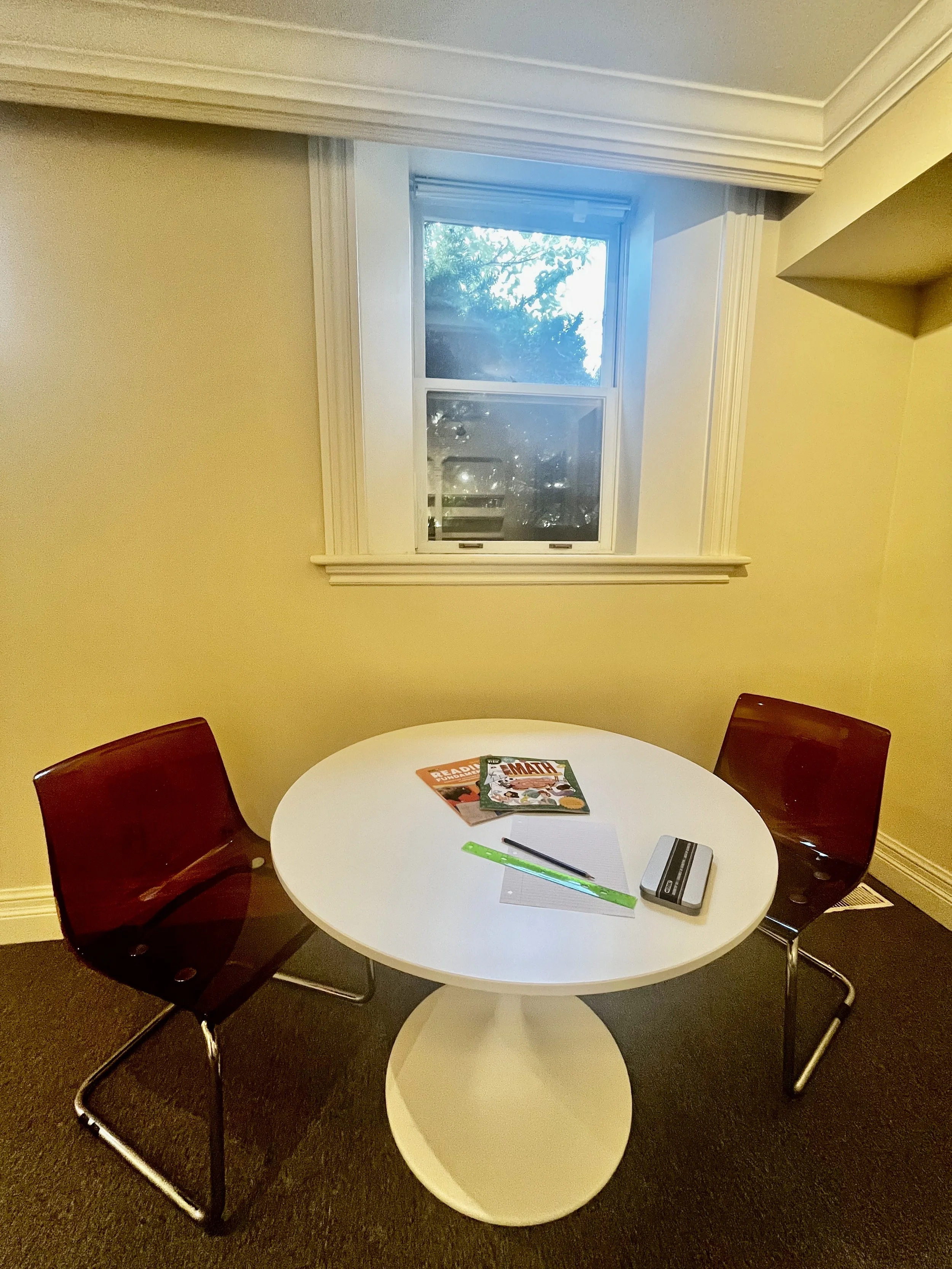A small round white table with two red chairs, books, a notepad, a pencil, a ruler, and a box on it, next to a window with a view of trees.