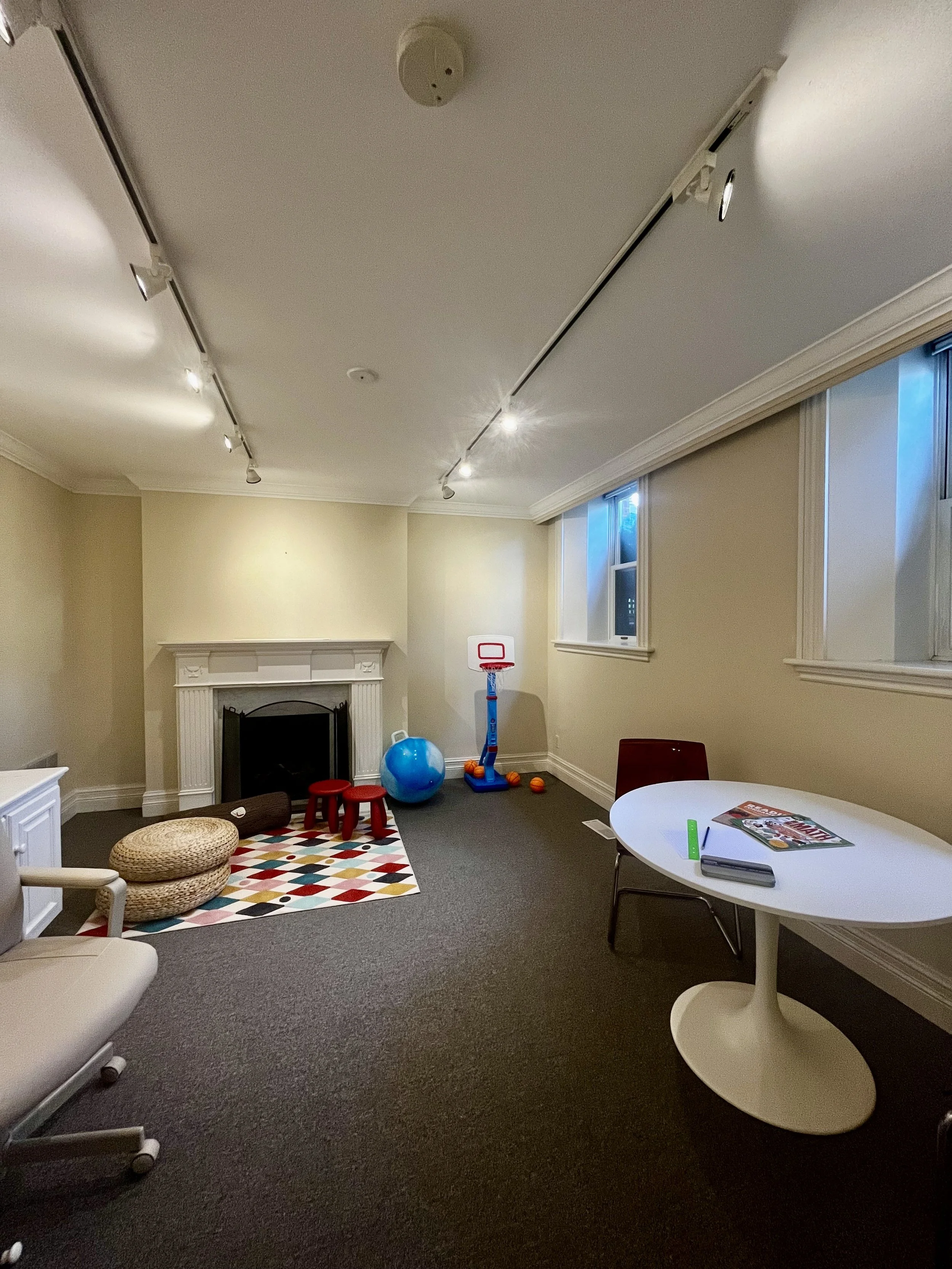 Room with beige walls, dark carpet, fireplace, small children’s play area with a colorful rug, ball, and basketball hoop, and a white round table with books and a ruler.