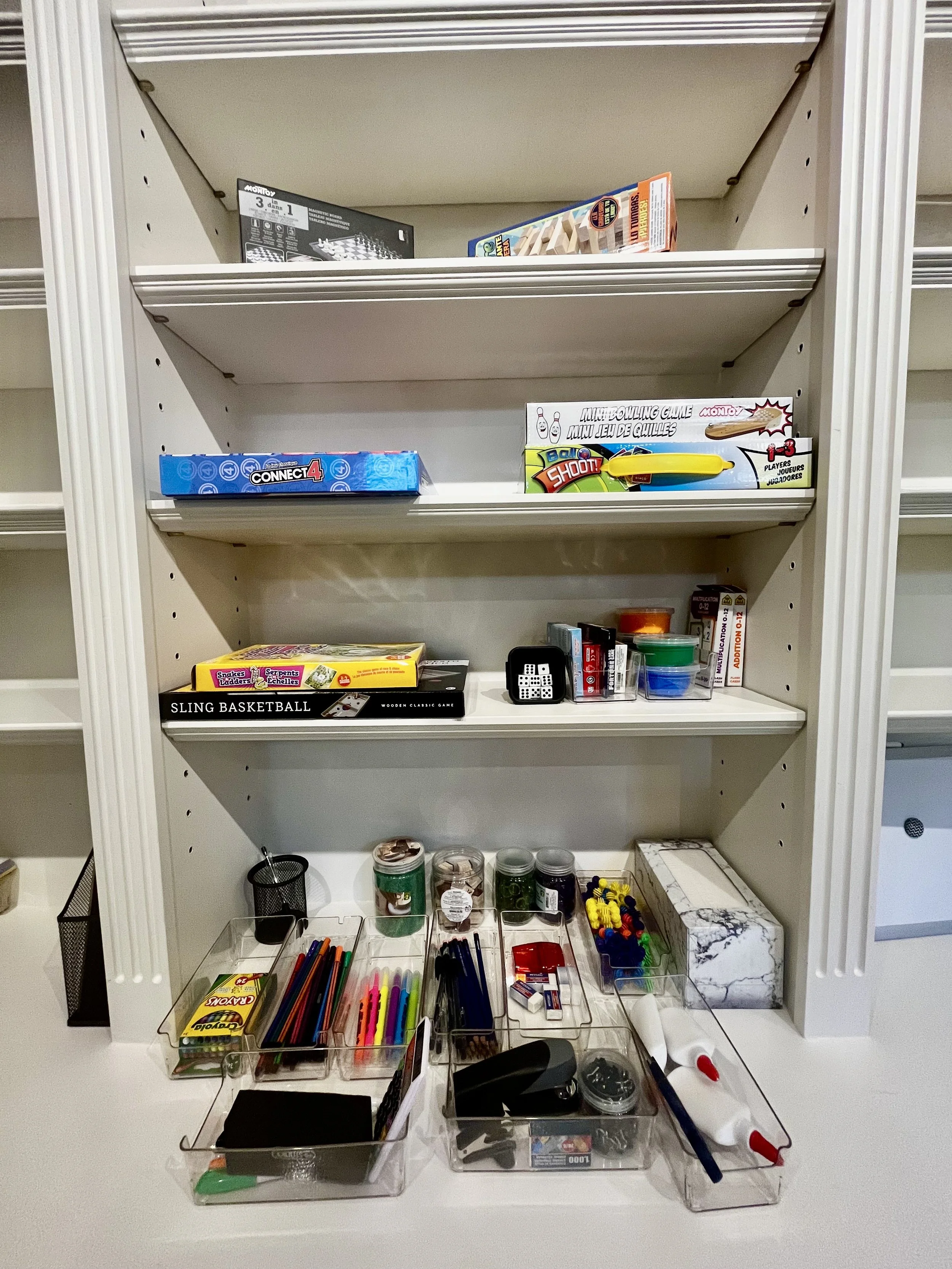 Organized bookshelf with board games, toy dice, and craft supplies on white shelves and clear bins.