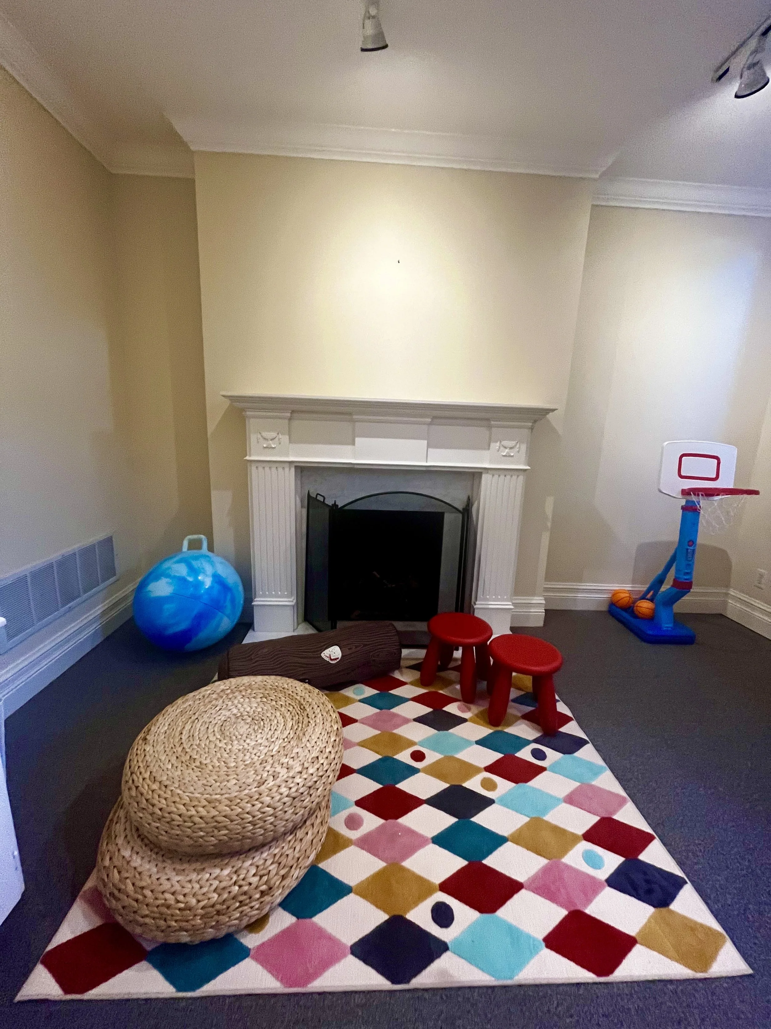 Class room with a colorful geometric rug, two red stools, a wicker ottoman, a blue exercise ball near a fireplace, and a small basketball hoop in the corner.