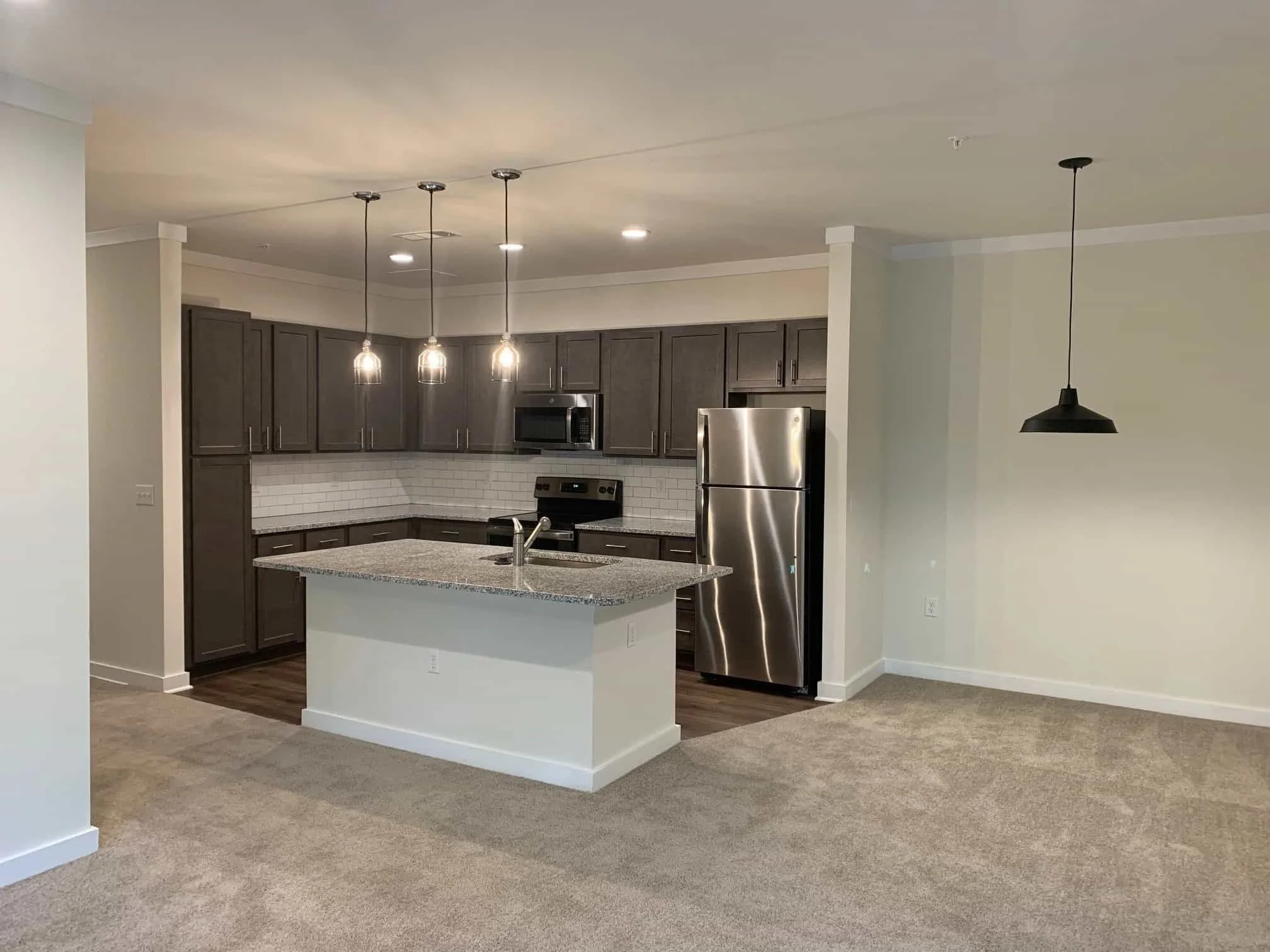 Modern kitchen with grey cabinets, stainless steel refrigerator, microwave, stove, granite countertops, white subway tile backsplash, island with sink, pendant lights, and open living space.