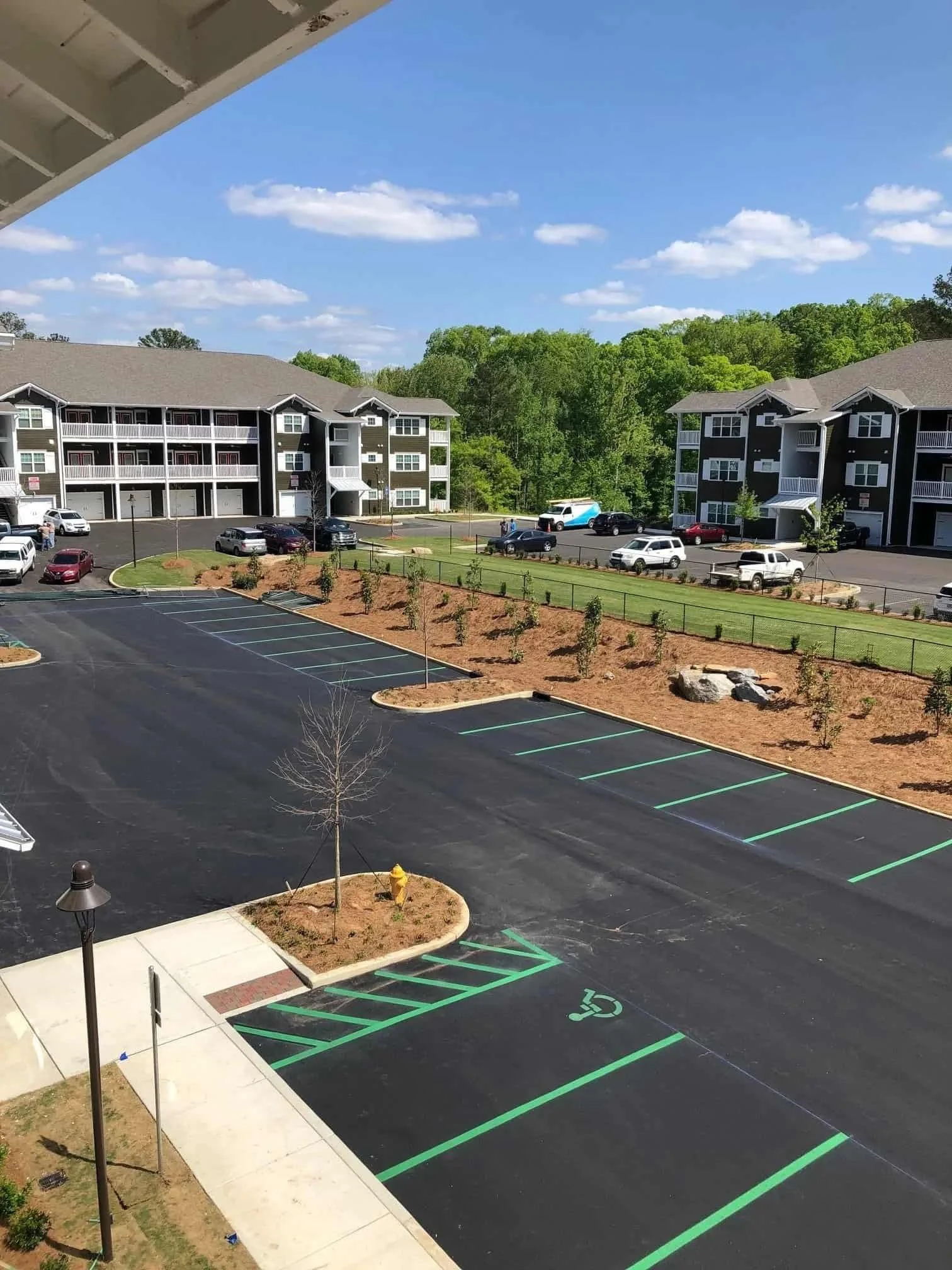 Empty parking lot with newly painted spaces, including a designated handicapped spot near a sidewalk with small trees and a fire hydrant, apartment buildings and cars in the background under a bright blue sky with scattered clouds.