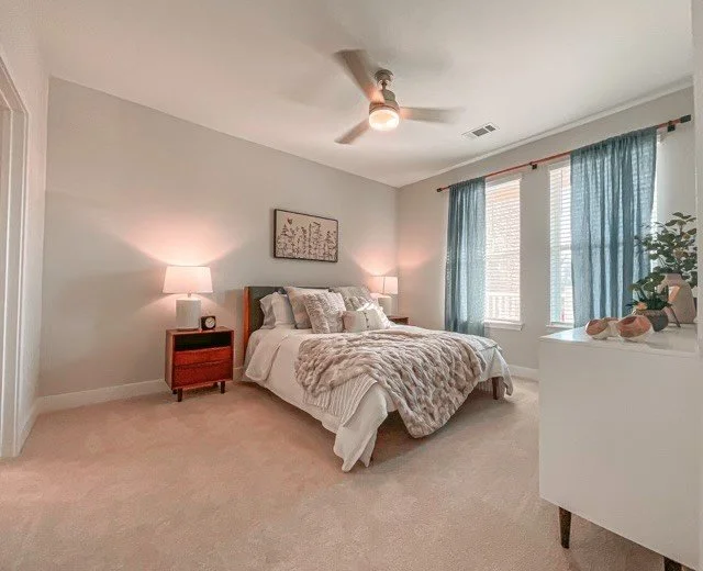 Bedroom with a large, sunny window, blue curtains and a patterned duvet within a typical unit in the Courtney Ridge multifamily apartment complex - Charlotte, NC