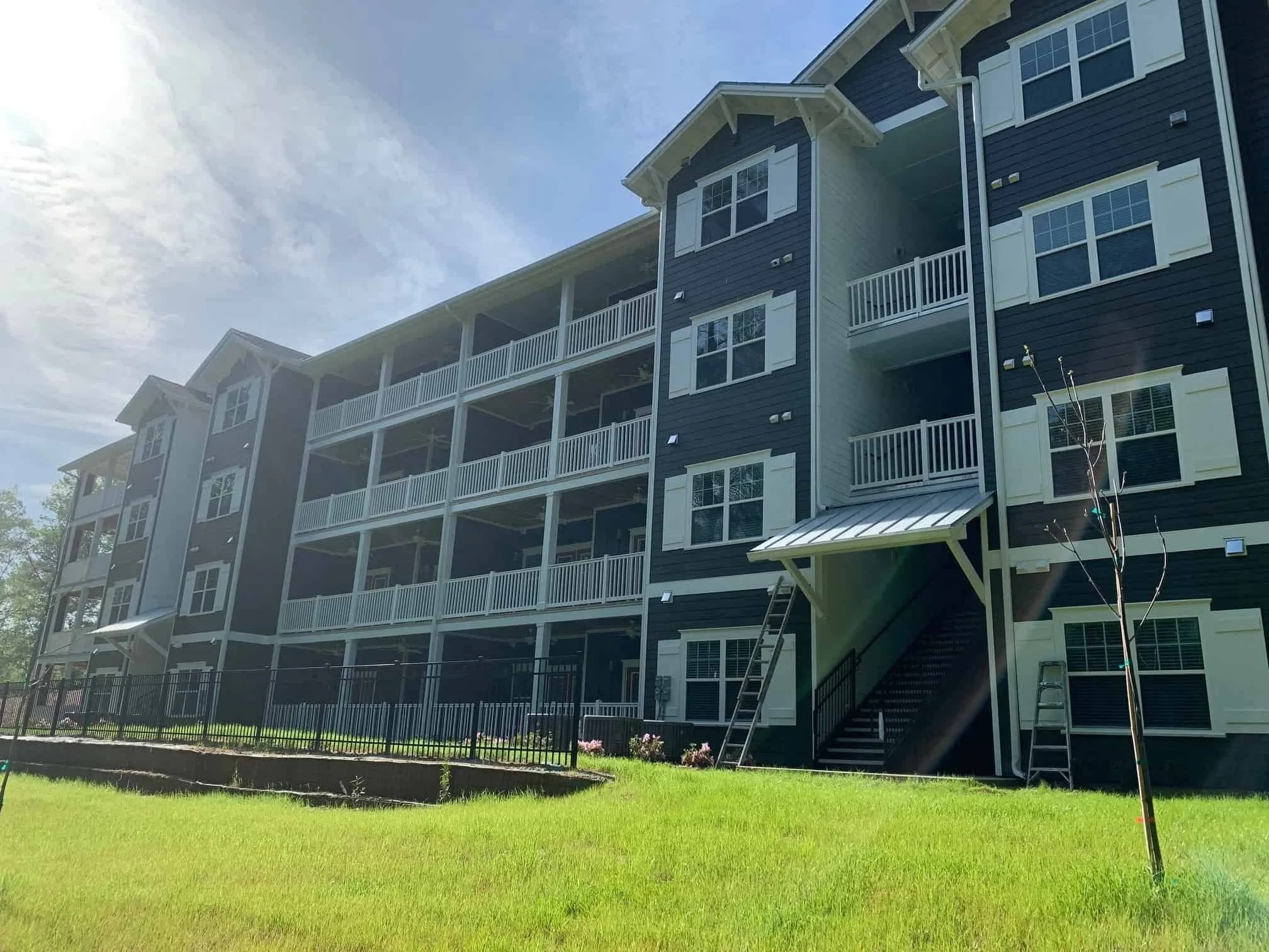 Multi-story residential building with dark siding and white window shutters, surrounded by green grass and a small tree.