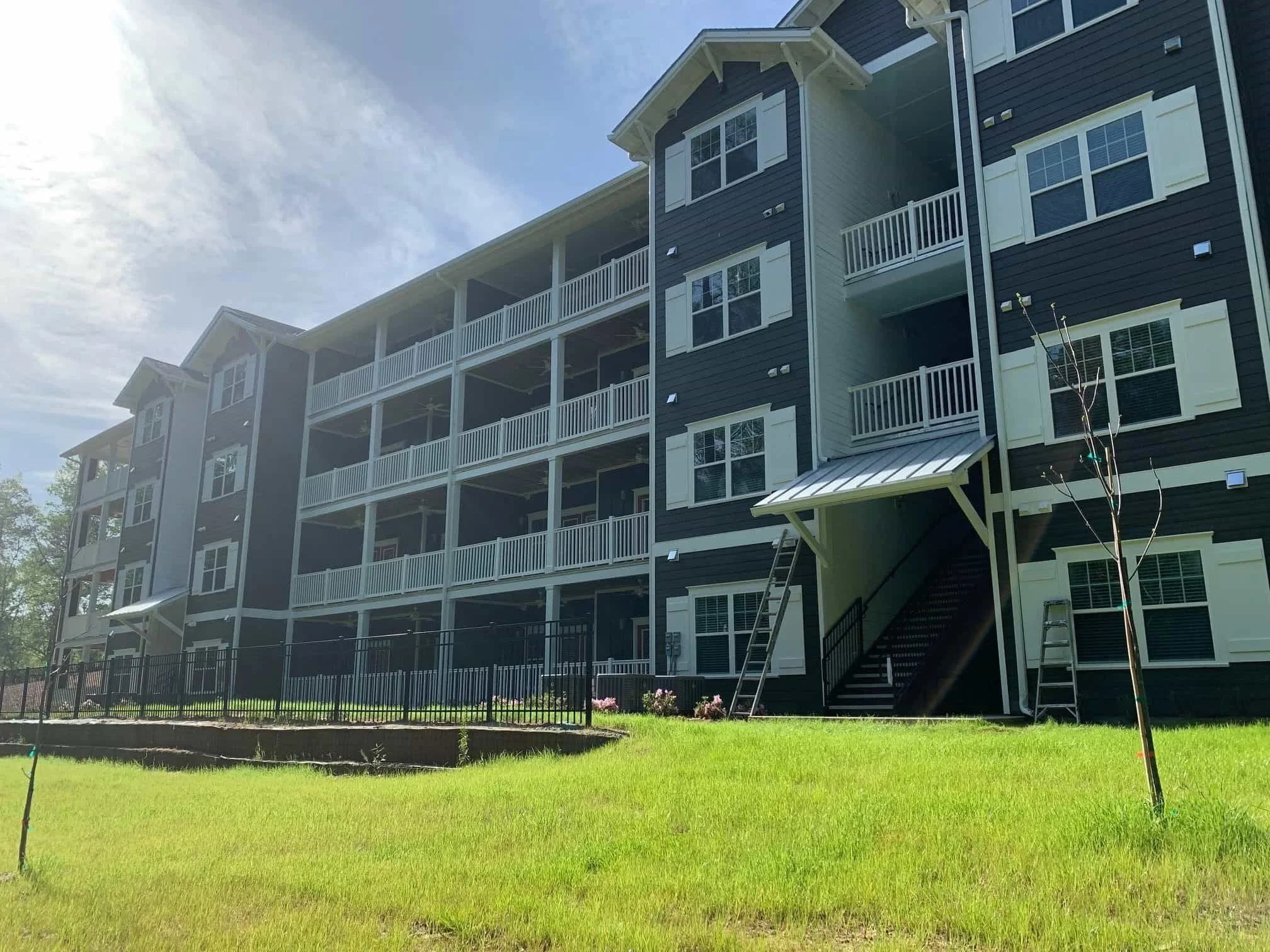 A multi-story residential apartment building with dark siding and white trim, surrounded by a green lawn, with small young trees and a staircase leading up to one of the entrances.