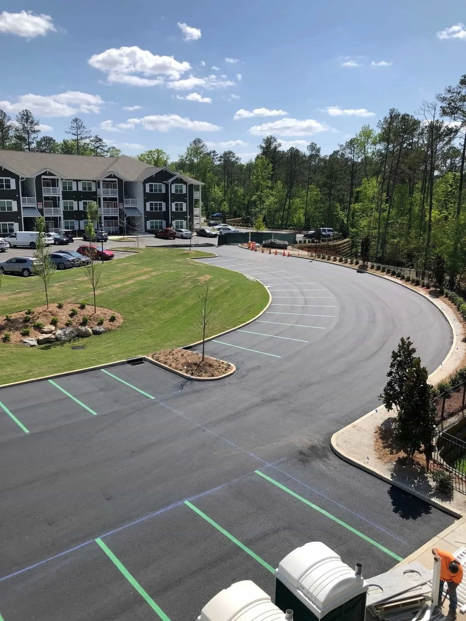 An empty parking lot with newly painted green lines, surrounded by trees and a grassy area, with a residential building in the background and a construction worker on the right.