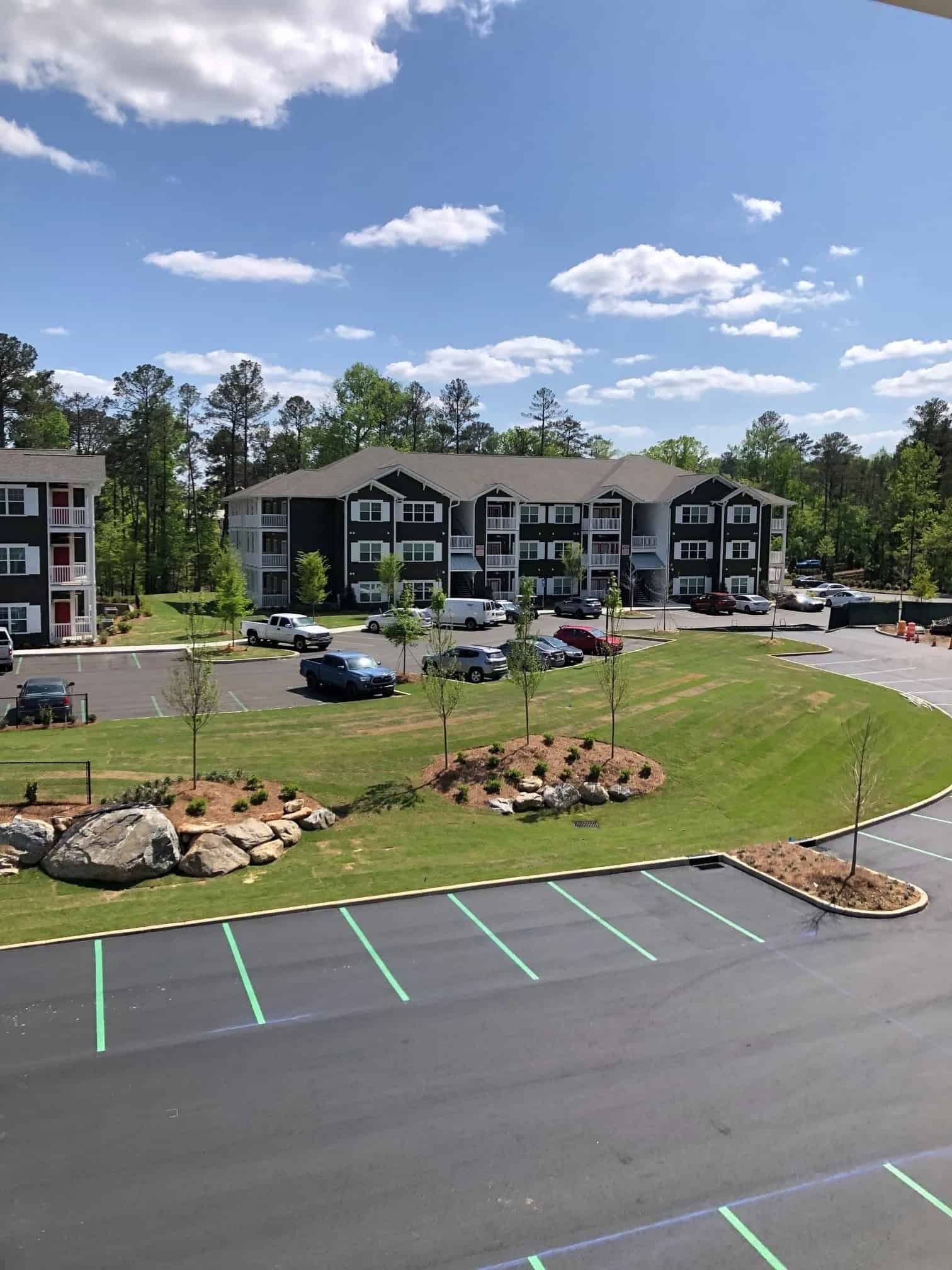 View of a parking lot with several striped parking spaces and landscaped areas featuring trees, rocks, and small plants, with a three-story apartment or condo building in the background under a blue sky with scattered clouds.