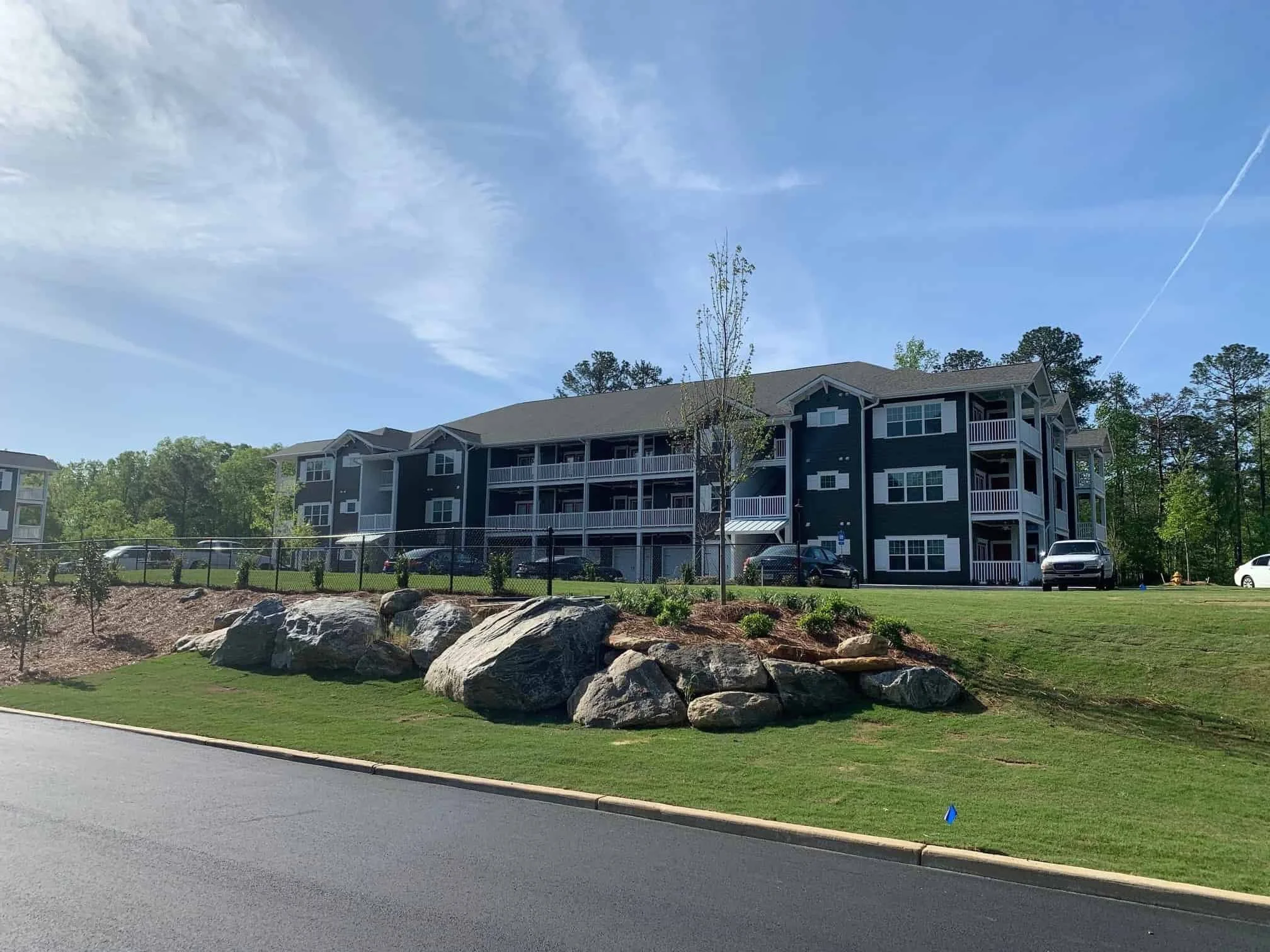 A multi-story residential building with balconies, parked cars in front, a grassy landscaped area with rocks, a young tree, and a clear blue sky.