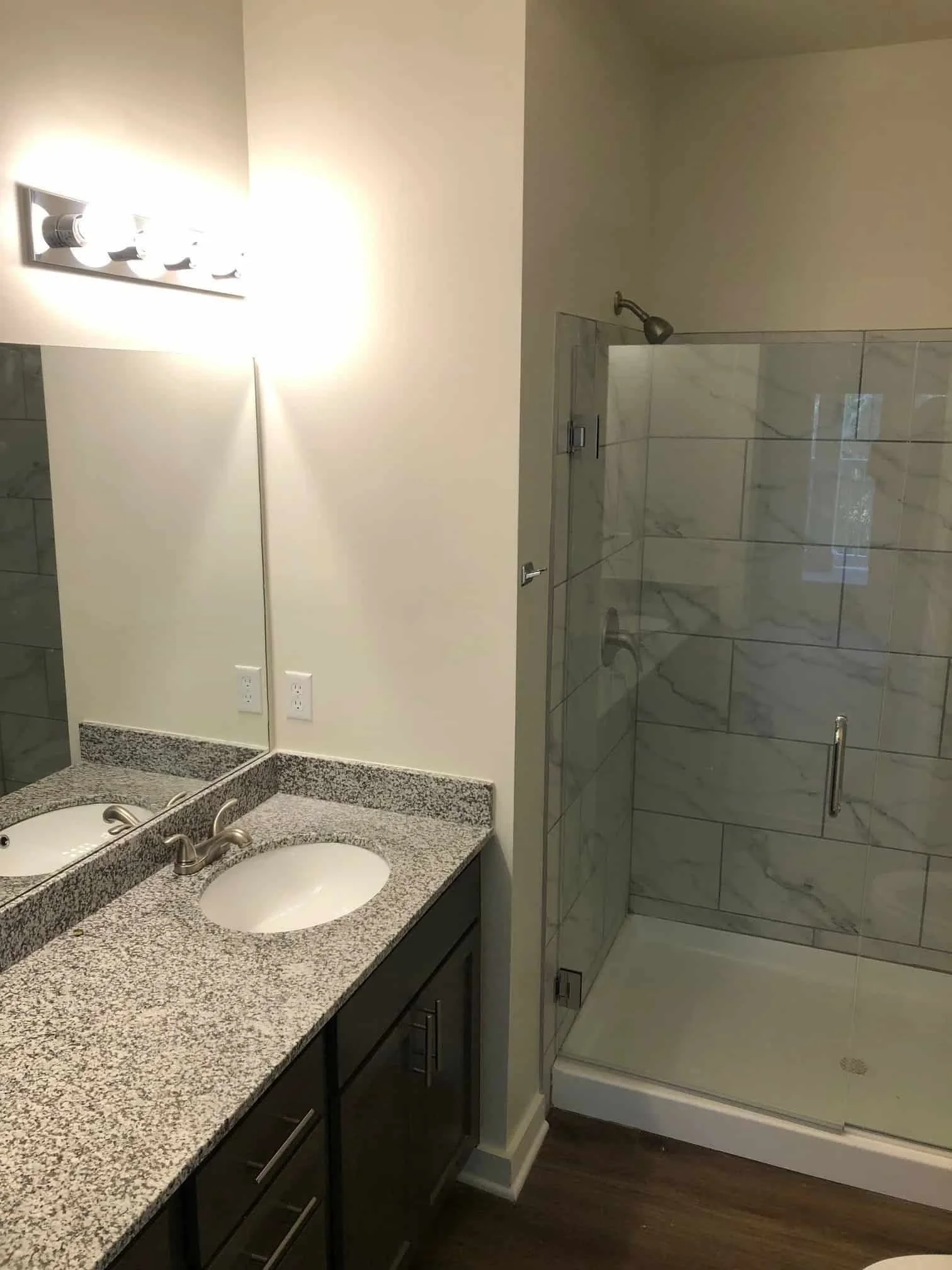 Bathroom with granite countertop vanity, round sink, mirror, wall-mounted lights, electrical outlets, and glass-enclosed shower with marble tiles.