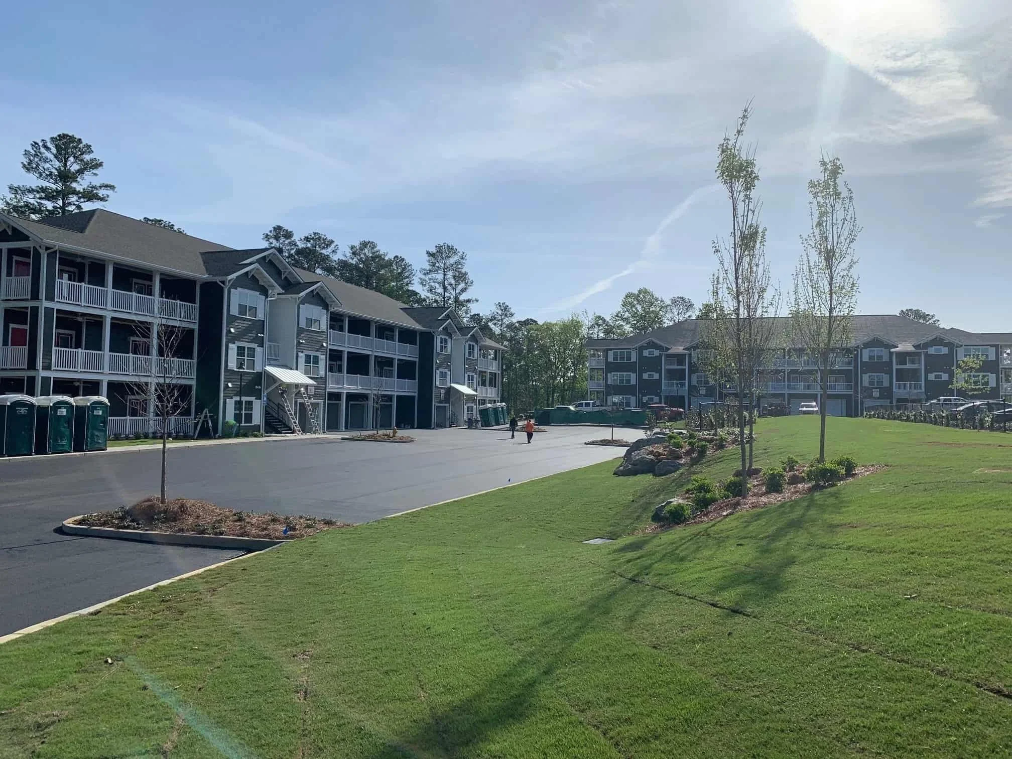 Apartment complex with multiple three-story buildings, a large paved parking area, and a well-maintained grassy lawn with young trees.
