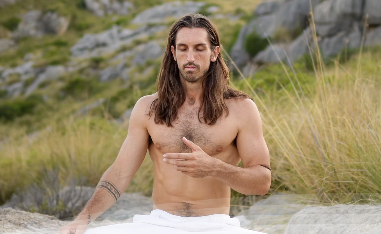 A shirtless man with long hair and tattoos practicing meditation outdoors in a grassy, rocky landscape.