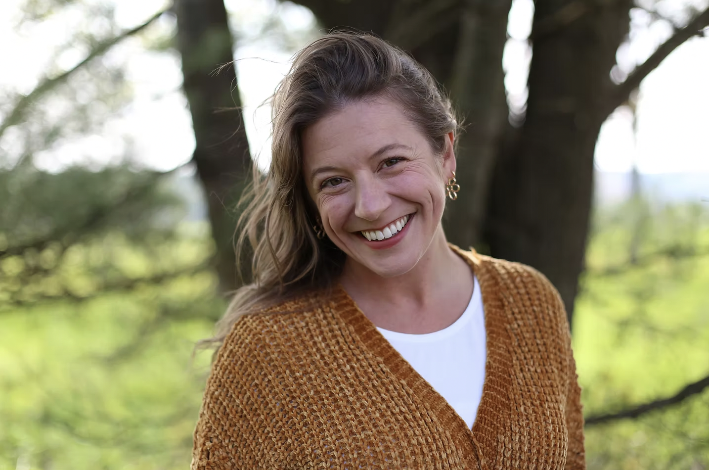 A woman with long wavy hair smiling outdoors in front of a tree, wearing a brown sweater and gold earrings.
