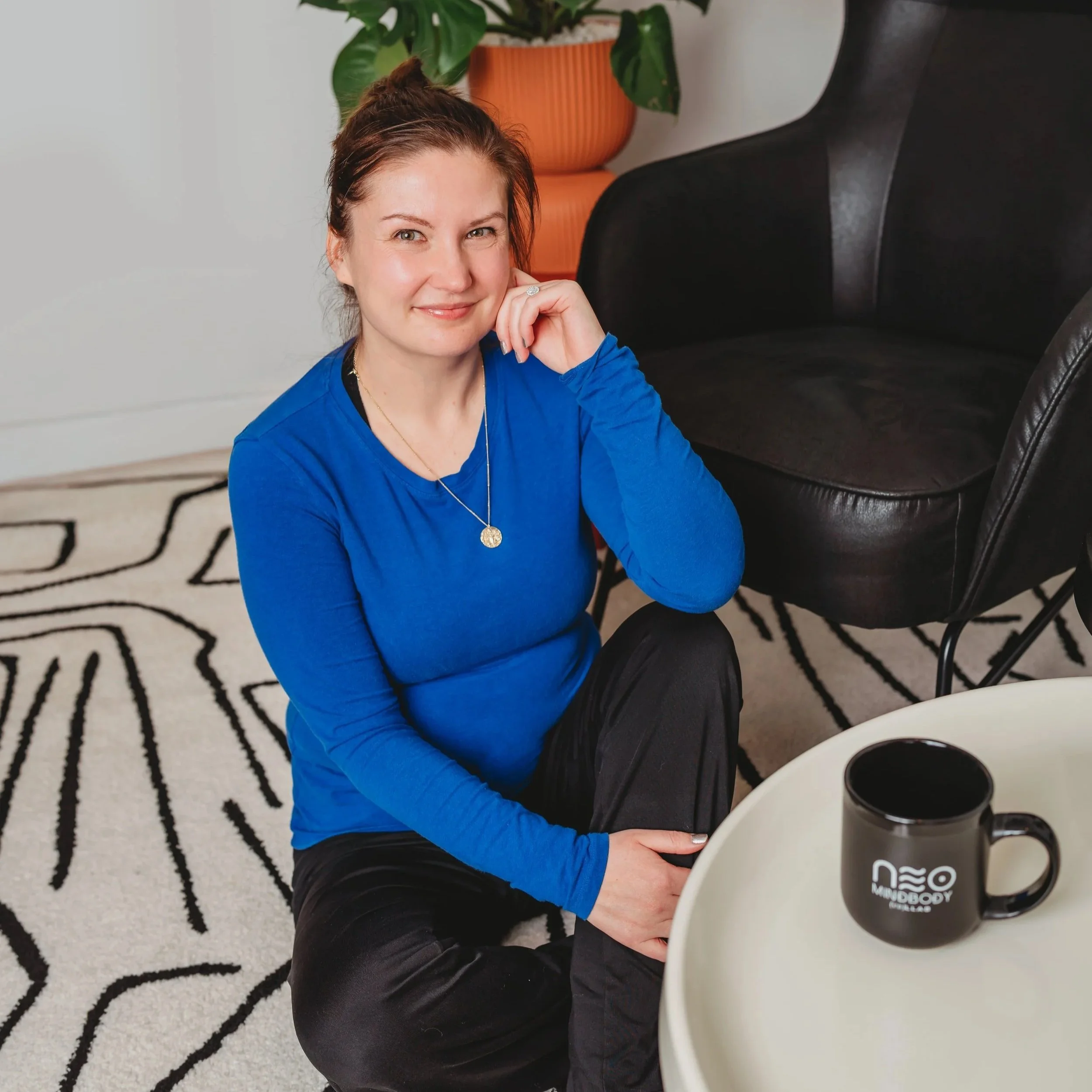 Woman with long dark hair and glasses sitting on a yoga mat with exercise equipment, including a black medicine ball, dumbbells, and a foam roller, in a room with a plant and contrasting black and white walls.