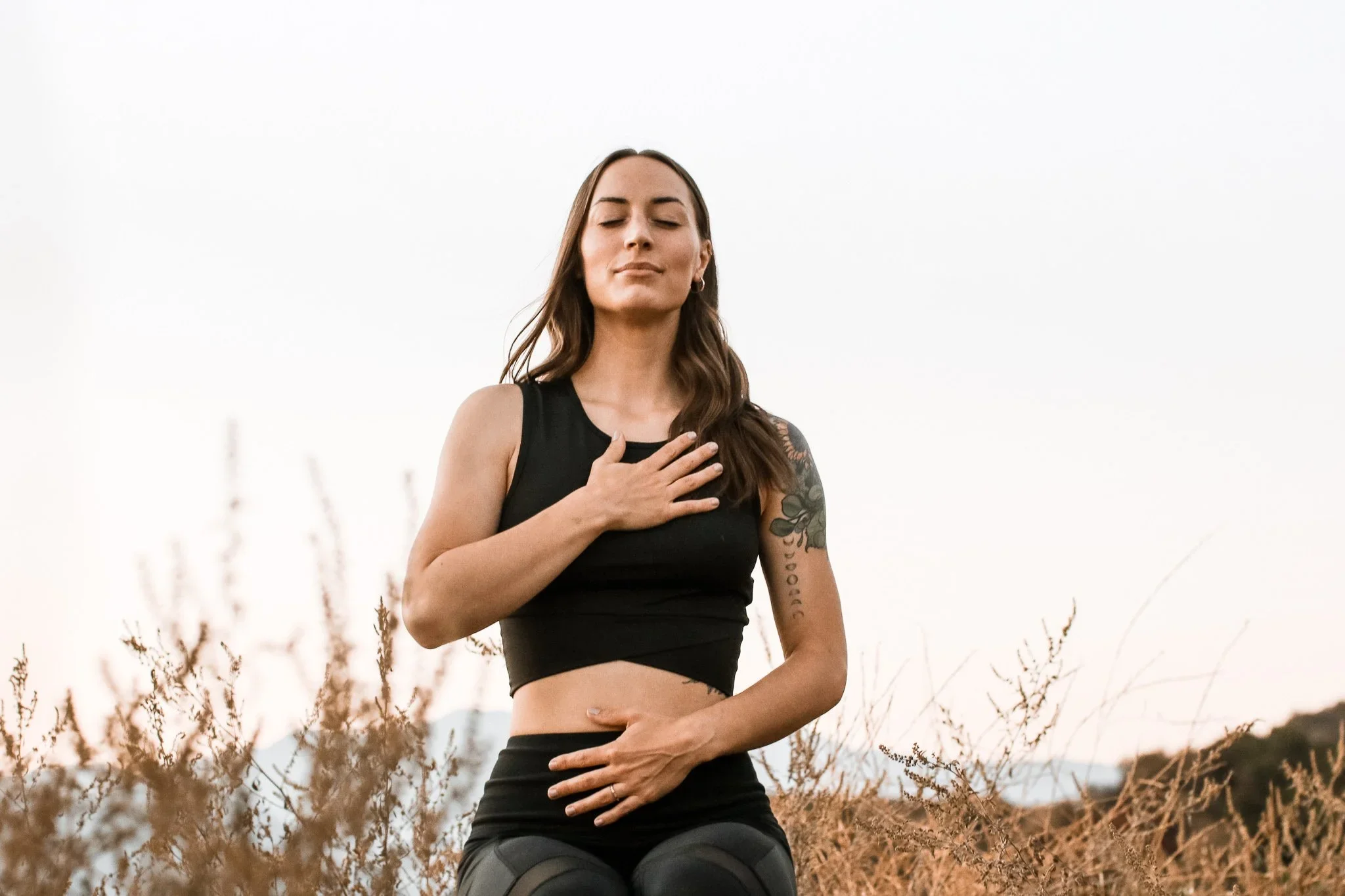 A woman with long brown hair, tattoos on her left arm, wearing black athletic clothing, is outdoors in a field with tall dry grass, with her hand on her chest and stomach, eyes closed and a peaceful expression.