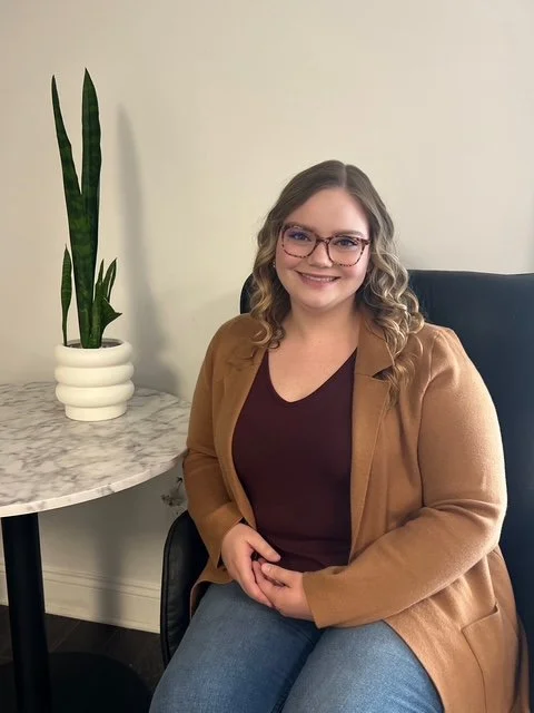 A woman with glasses and wavy blonde hair sitting on a black chair, smiling, wearing a brown blazer and a maroon top, next to a marble-topped table with a tall, green snake plant in a white pot.