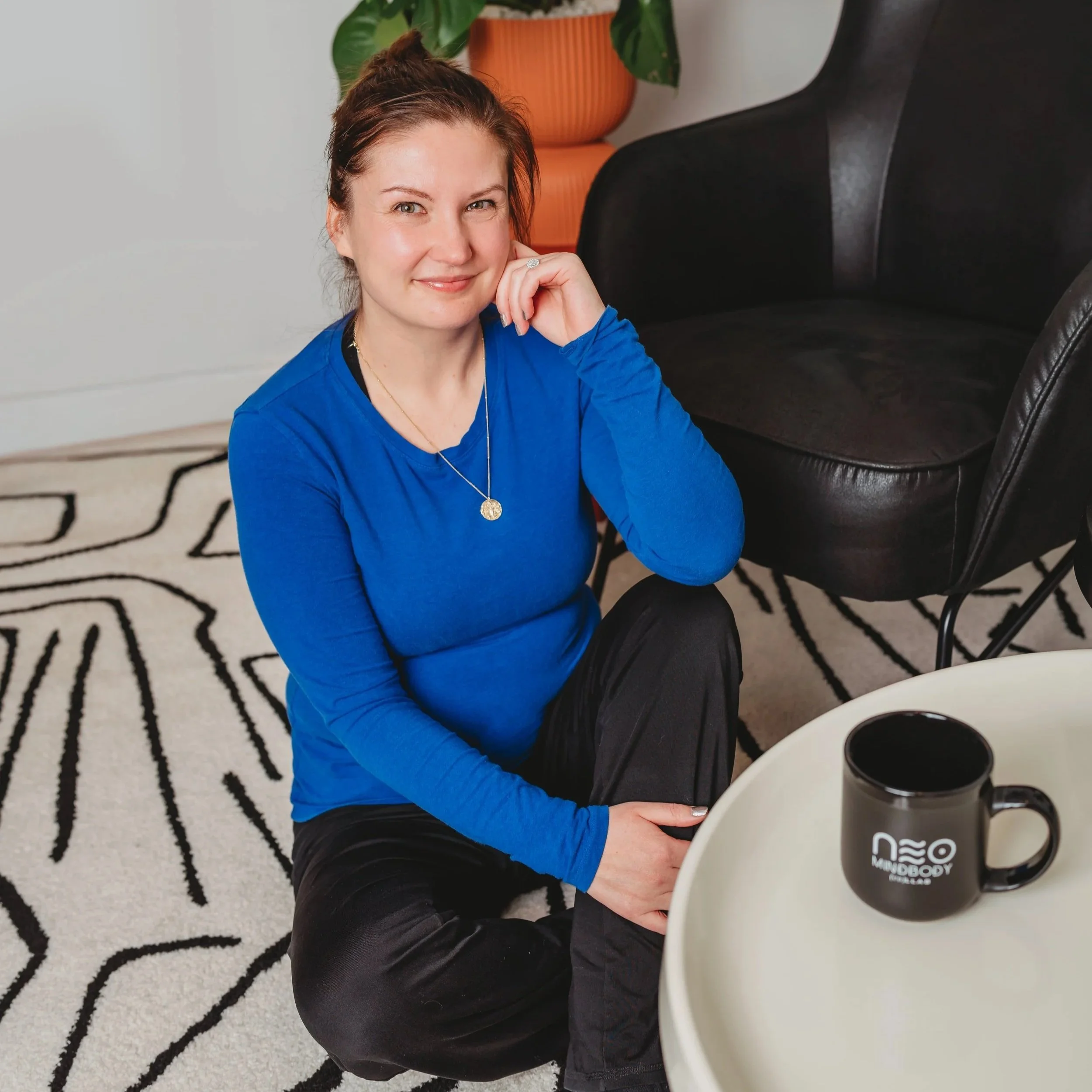A smiling woman with long blonde hair in a red polka dot blouse sitting on a tan leather bench against a black and white wall.