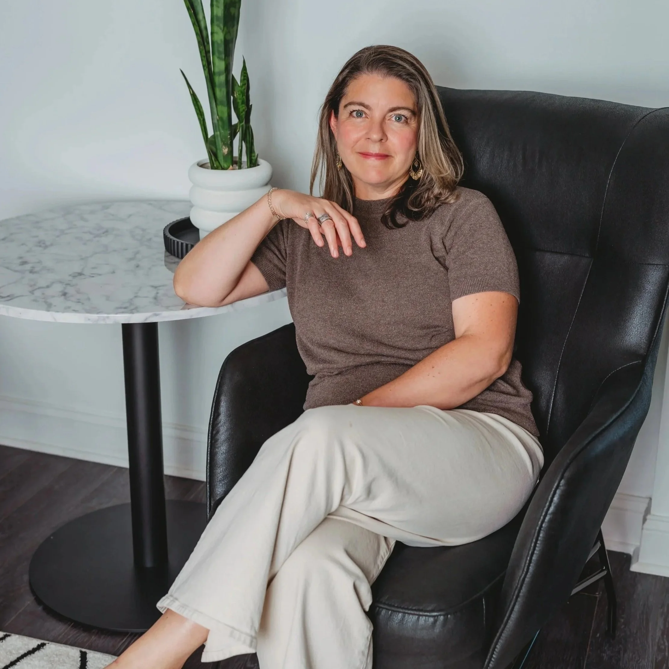 A woman sitting in a black leather armchair near a round marble-top table with a potted snake plant, with a white wall in the background.